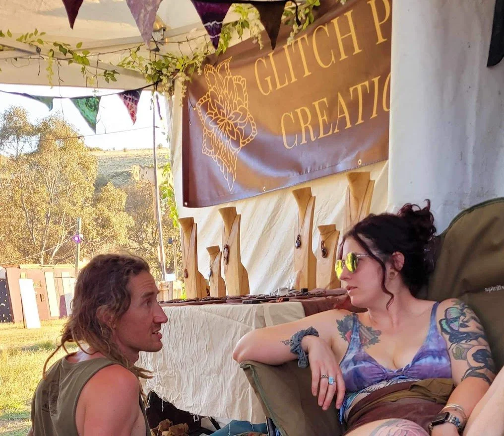 Two women at an outdoor festival or event, sitting and talking. One woman with long hair is sitting on the ground, and the other with dark hair and tattoos is reclining in a chair wearing sunglasses. There is a booth or display with a banner that say