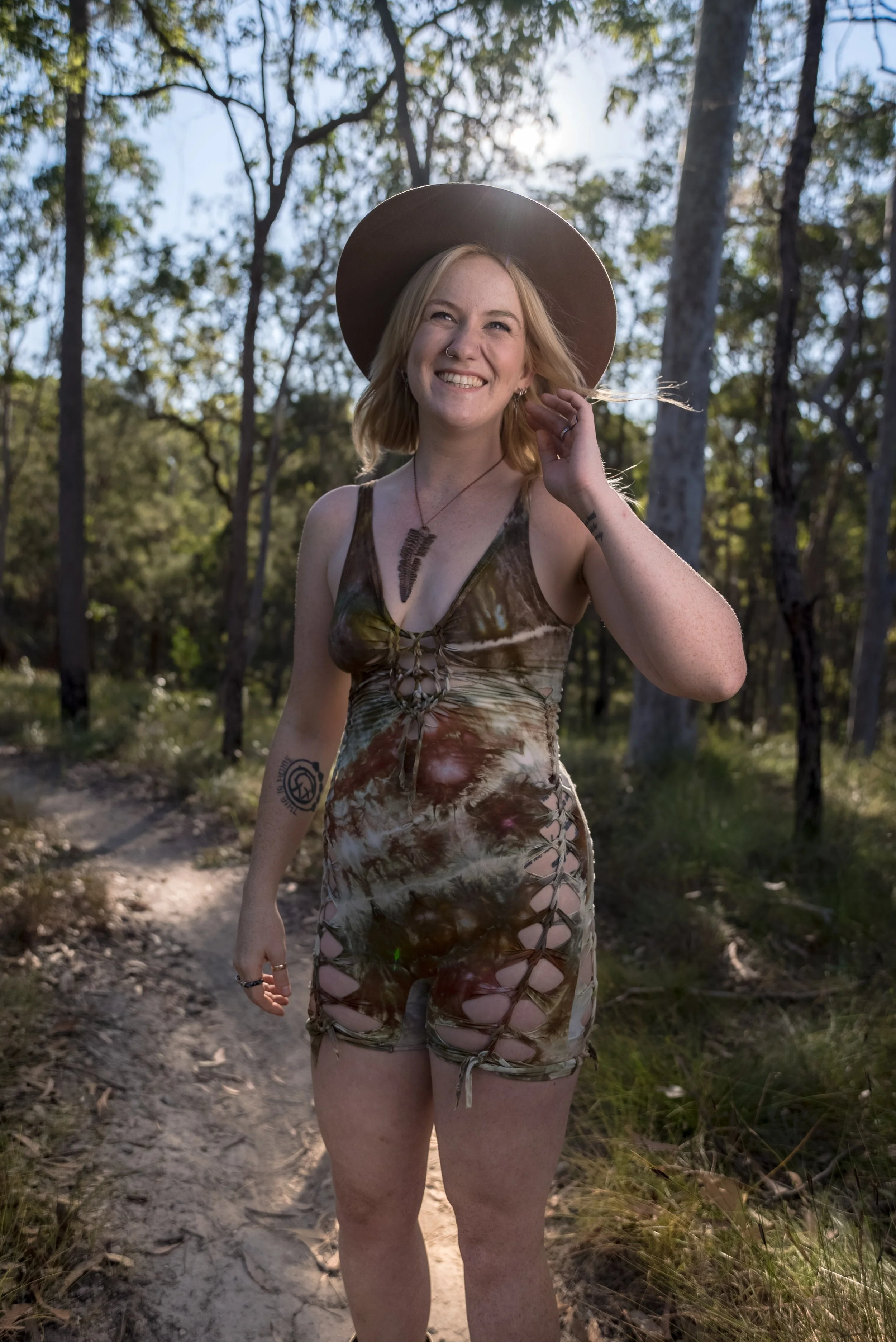 A smiling woman with blonde hair in a wide-brimmed hat standing on a dirt path in a wooded area during daylight.