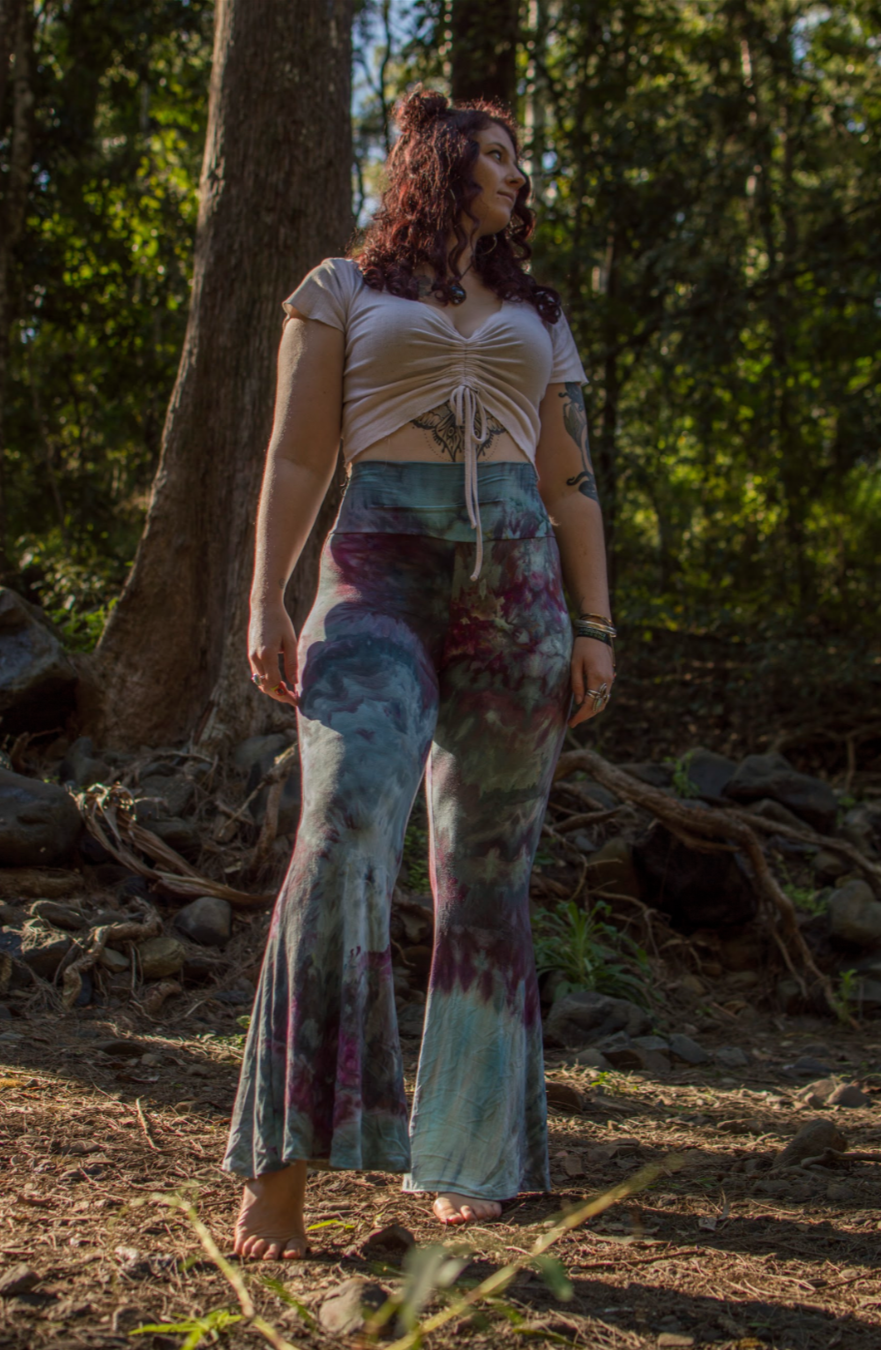 A woman with curly hair standing barefoot in a forest, wearing a beige crop top and colorful tie-dye pants.