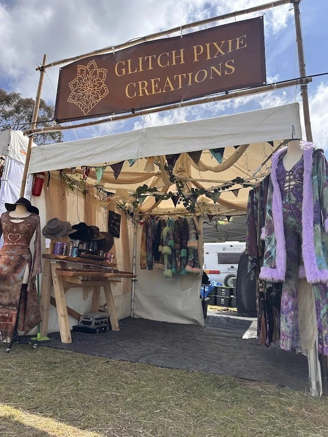 A booth at an outdoor craft fair with a sign reading 'Glitch Pixie Creations'. The booth displays tie-dye clothing, hats, and mannequins wearing dresses and jackets, with a cloudy sky overhead.