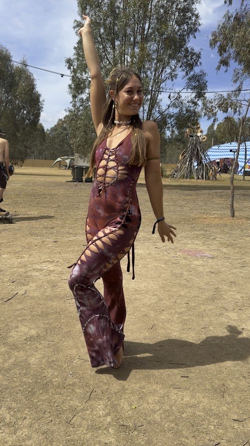 A young woman is dancing outdoors in a tie-dye jumpsuit with cut-out designs, raising her right arm and smiling. There are trees, tents, and other people in the background on a sunny day.