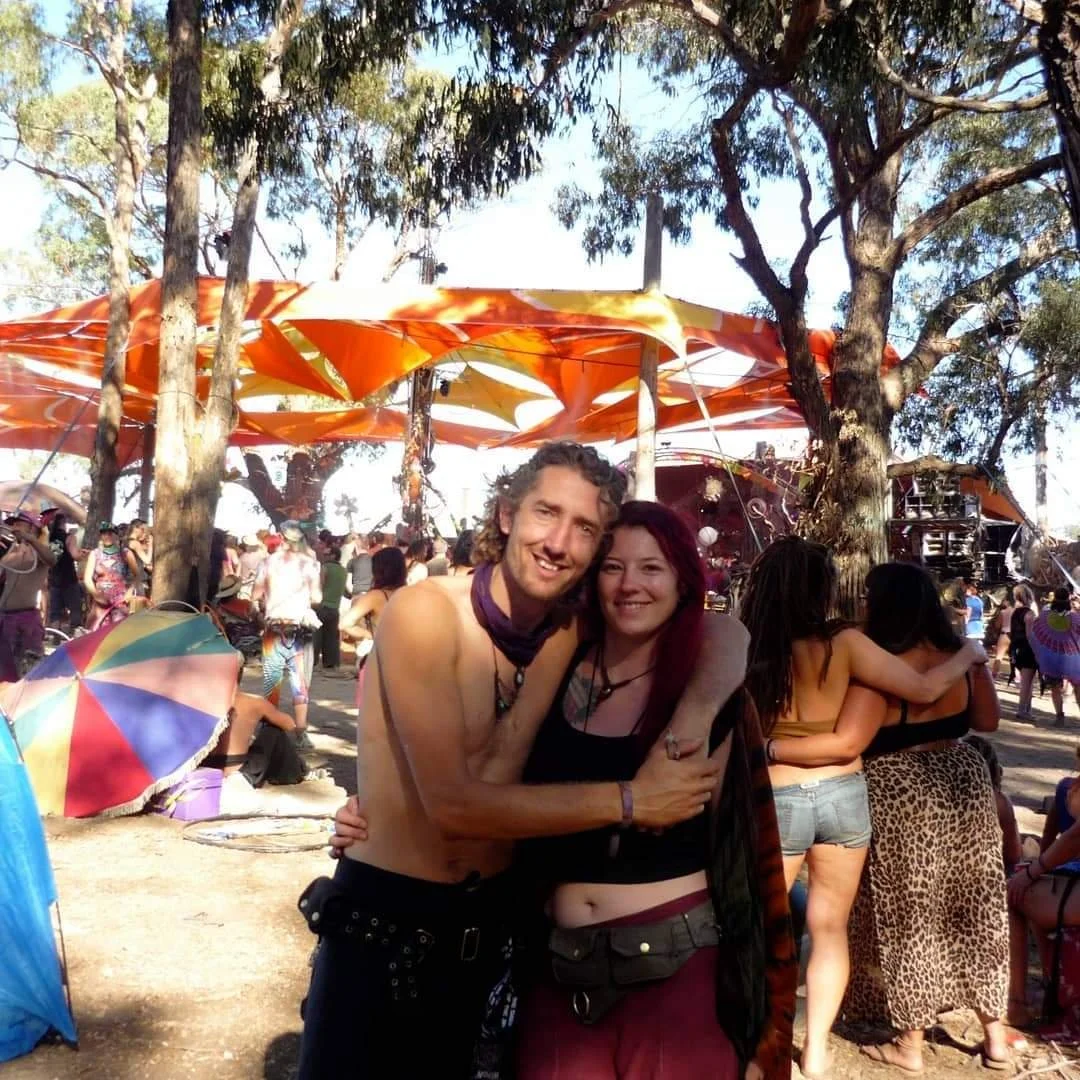 A man and woman smiling and hugging at an outdoor music festival, with a crowd and colorful umbrellas in the background.