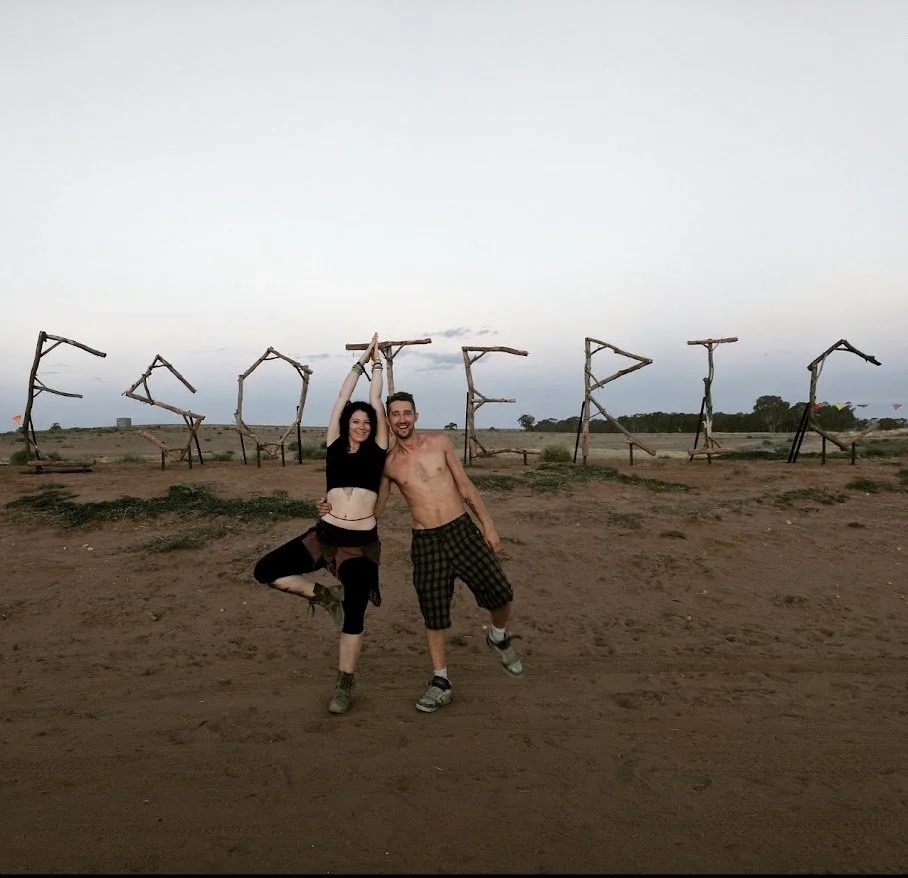 Two people, a woman and a man, dancing and smiling in an outdoor field during sunset, with the word 'SPROSTERS' spelled out in large wooden letters in the background.