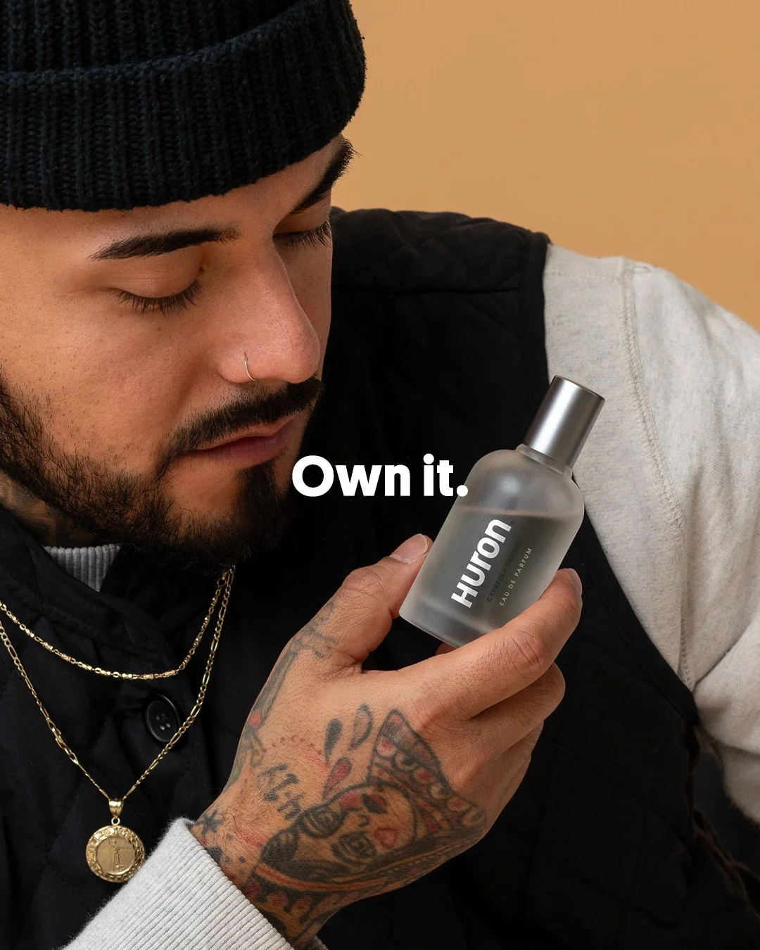 Close-up editorial studio portrait of a tattooed male athlete in a black beanie and vest looking at a Huron fragrance bottle.