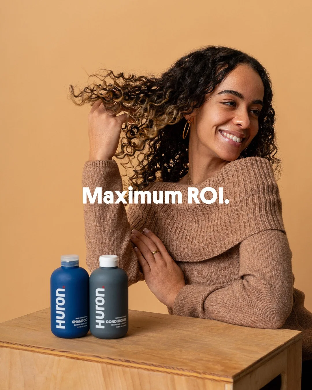 Editorial studio portrait of a smiling female athlete with curly hair sitting with Huron shampoo and conditioner bottles for the grooming result state.