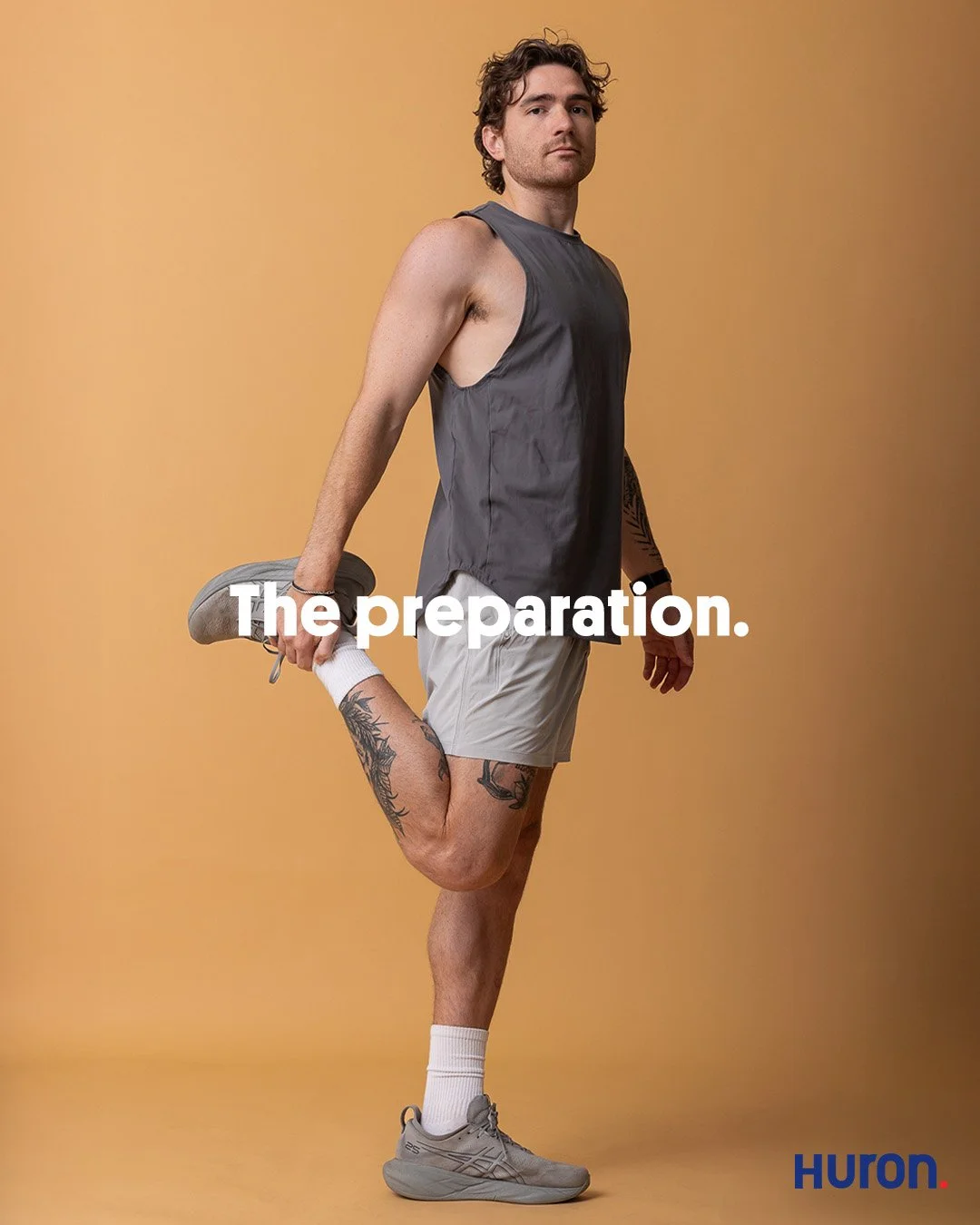 Full length studio portrait of a male runner with leg tattoos in a grey tank top stretching his quad against a tan background.