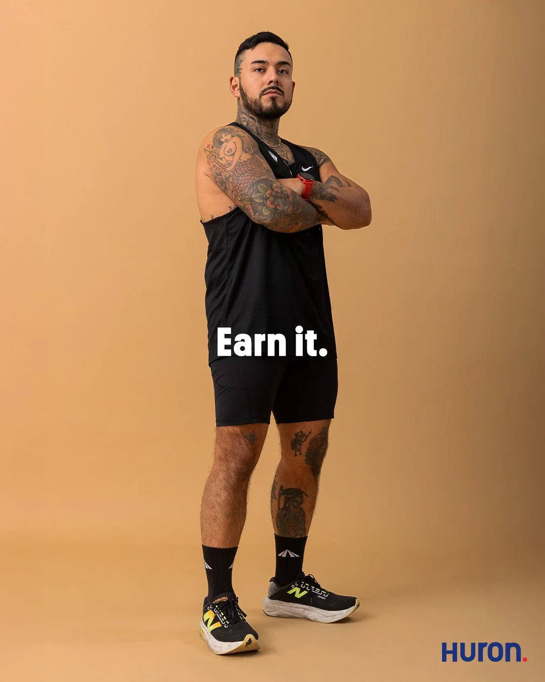 Studio portrait of a male athlete with extensive arm and neck tattoos standing with arms crossed against a tan background for the men's grooming brand Huron.