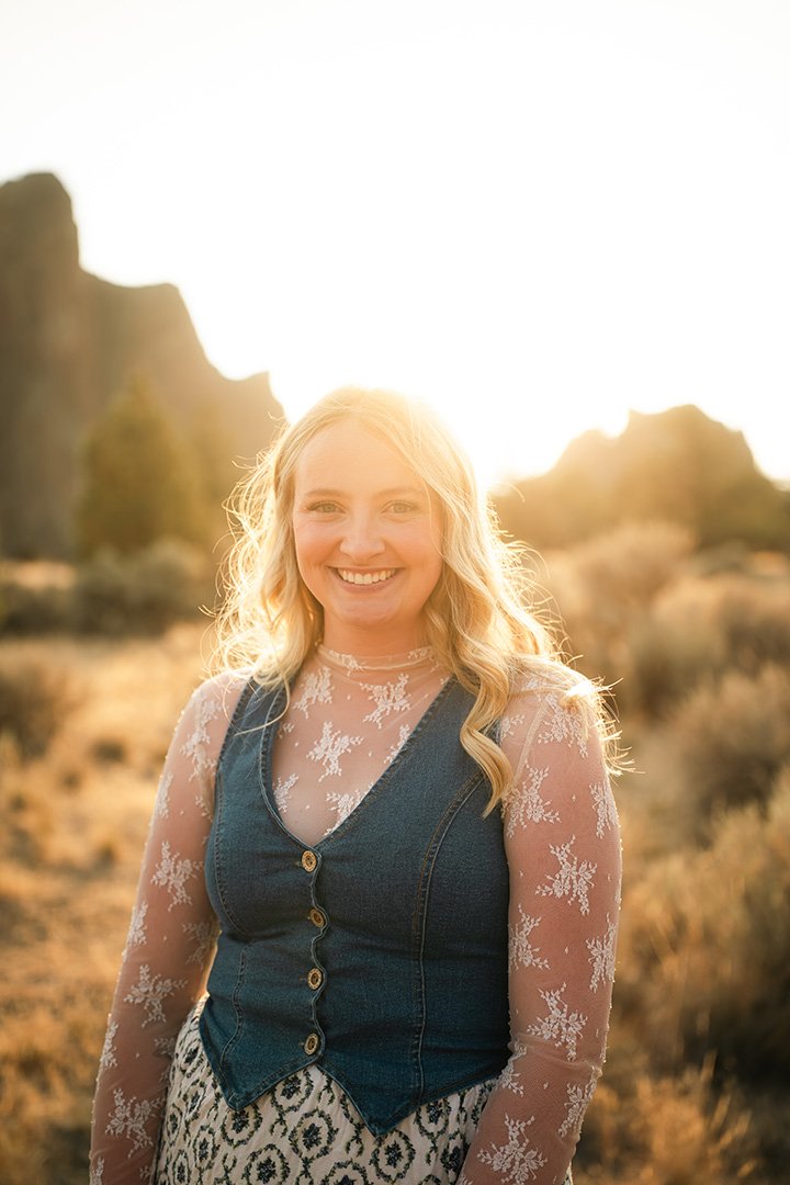 A smiling woman with blonde hair standing outdoors at sunset in a desert landscape.