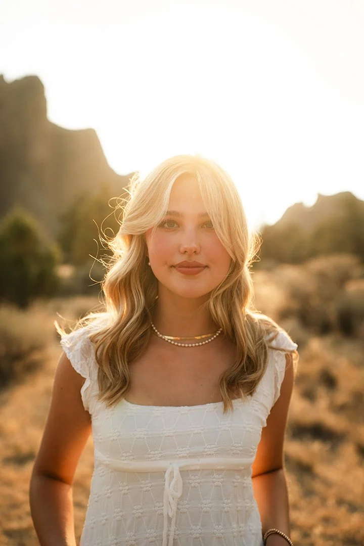 A young woman with blonde hair wearing a white dress and pearl necklace standing outdoors during sunset in a rocky, desert-like landscape.