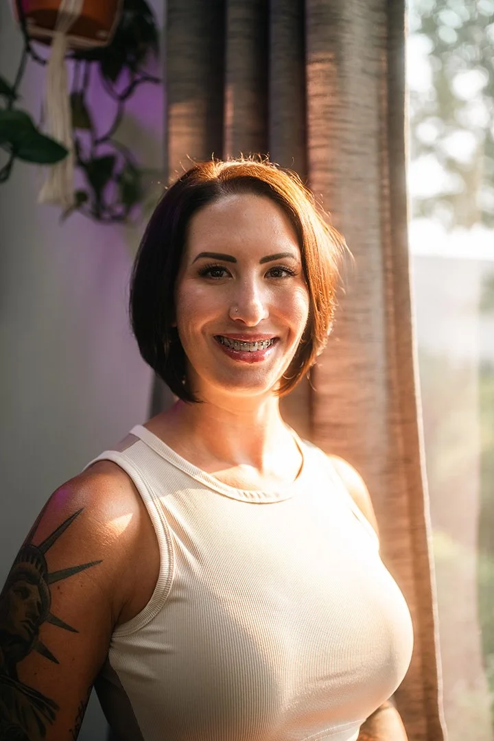 A woman with short dark hair and tattoos on her arm, smiling with braces, standing near a window with sunlight coming through, wearing a white sleeveless top.