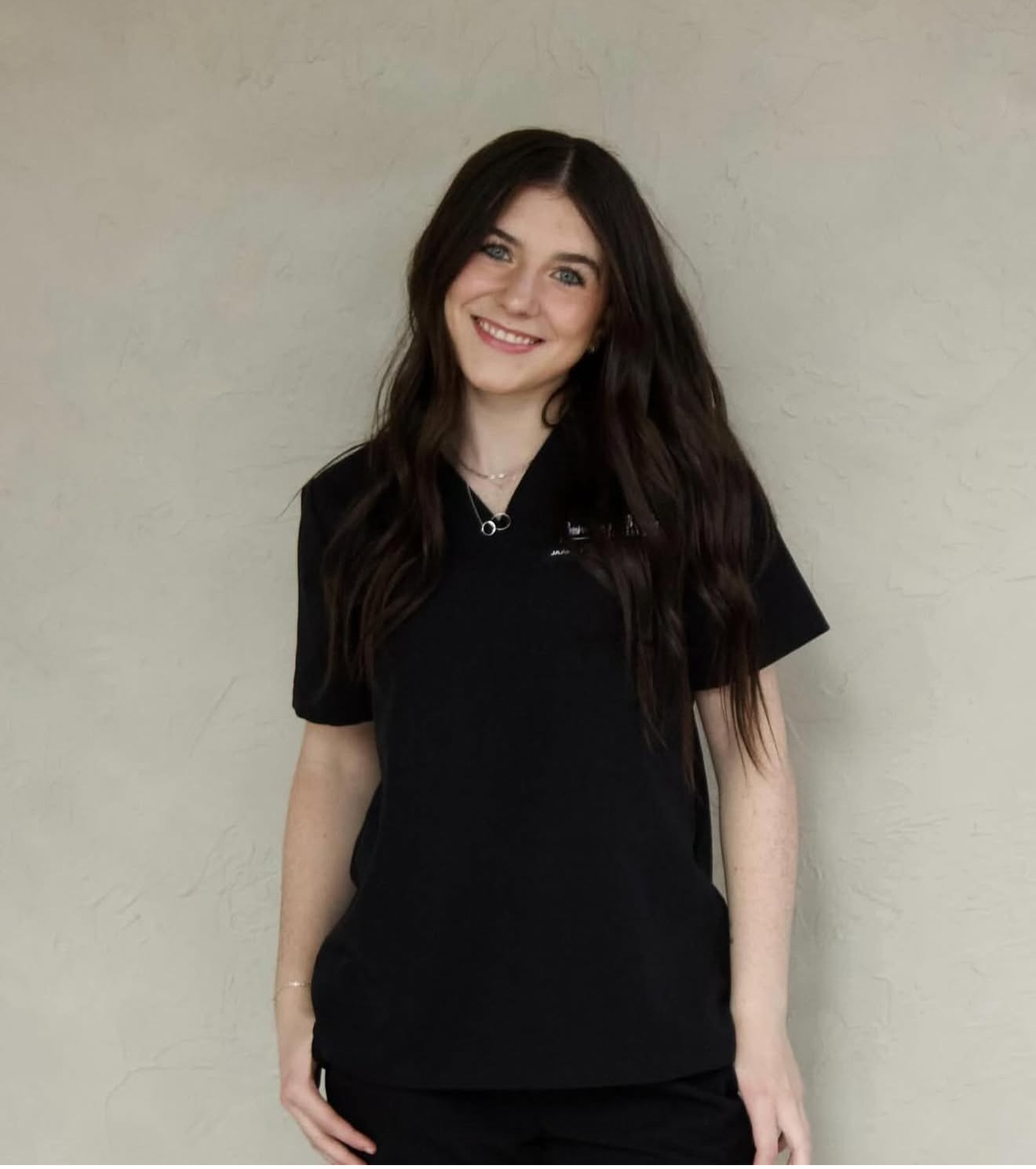 A young woman with long brown wavy hair, wearing a black t-shirt, standing against a plain beige wall and smiling at the camera.