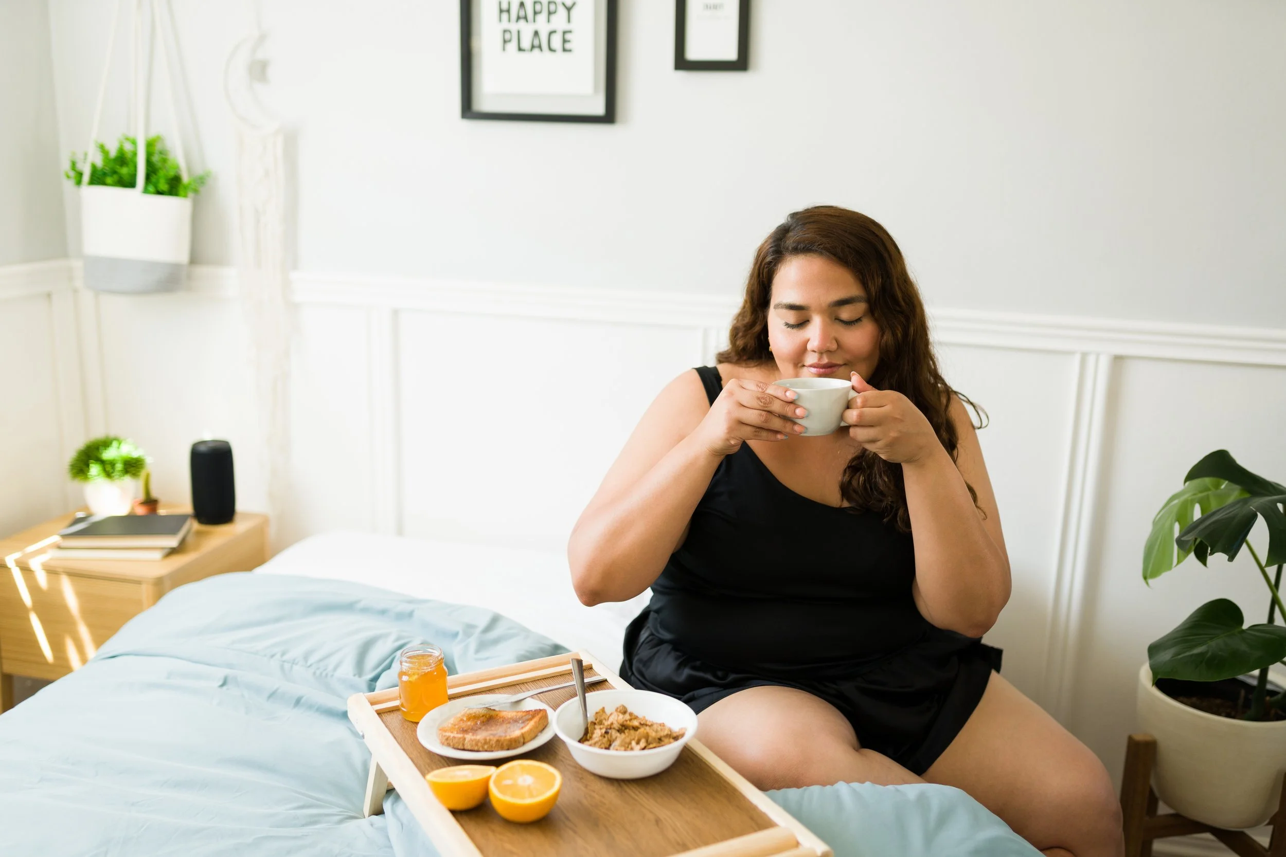 A woman of color enjoying coffee and breakfast in bed, symbolizing rest and caring for her own needs.
