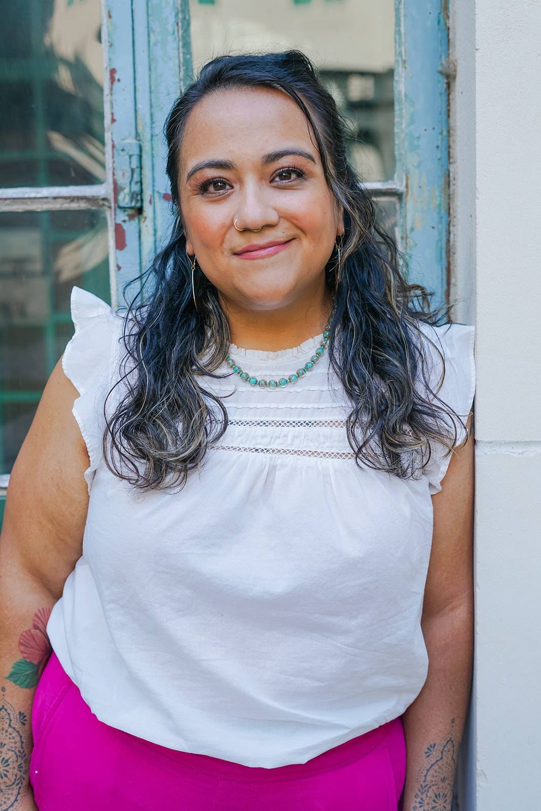 Monica Jurado Kelly, LCSW-S, therapist in Texas and Virginia, smiling in front of a teal window frame, wearing a white blouse and bright pink pants.