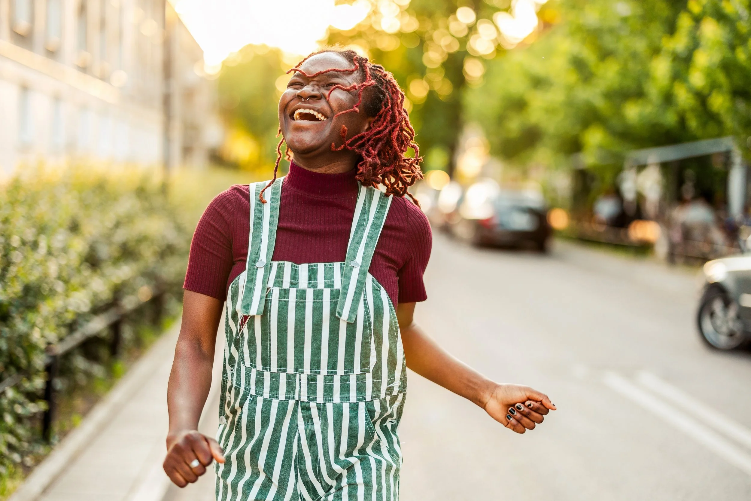 Black queer person with locs laughing joyfully outdoors in sunshine, arms open expressing freedom, relief, and joy after choosing rest over productivity