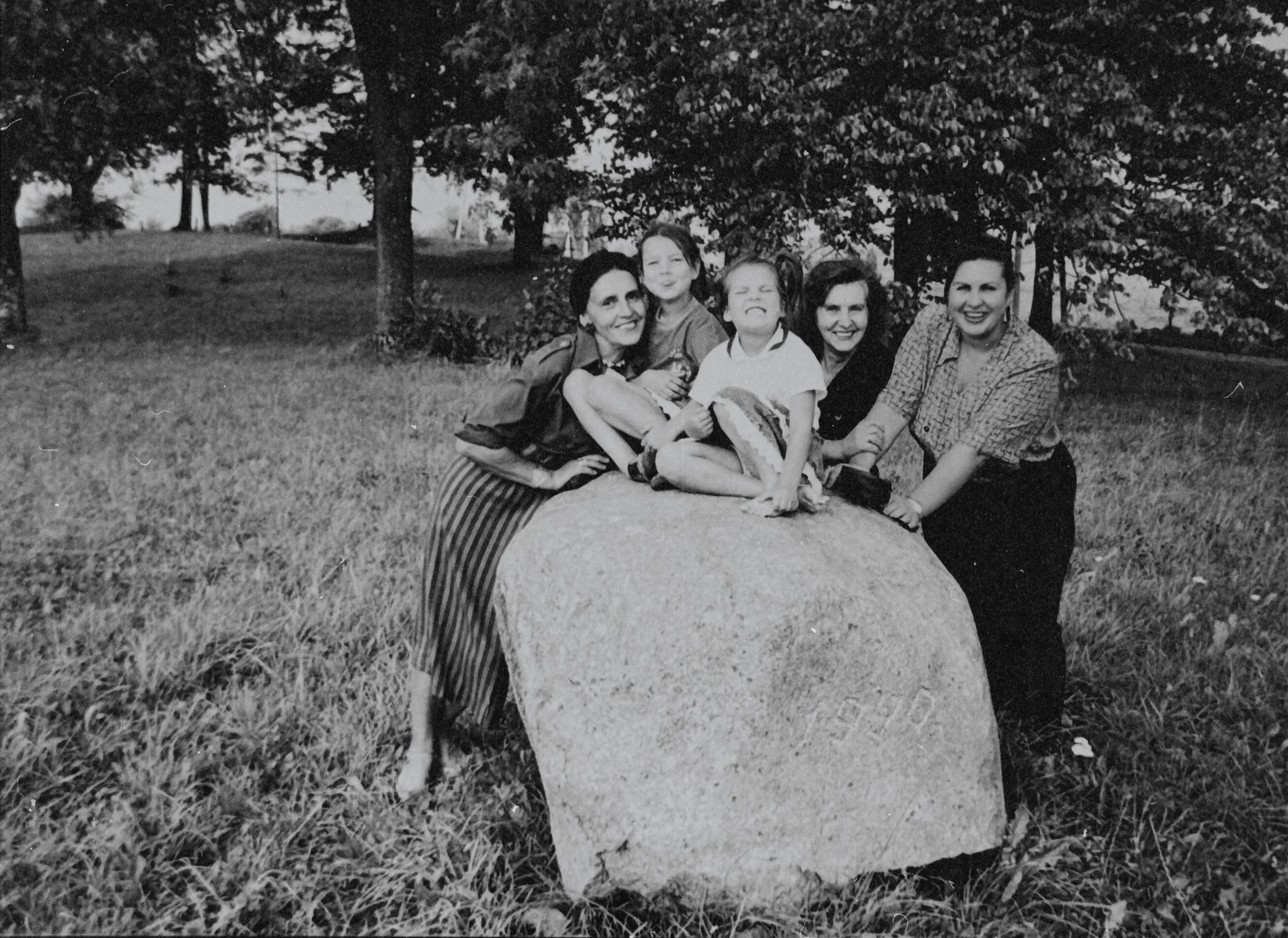 Black-and-white family photograph showing three women and two children laughing together outdoors, leaning on a large stone in a field surrounded by trees.