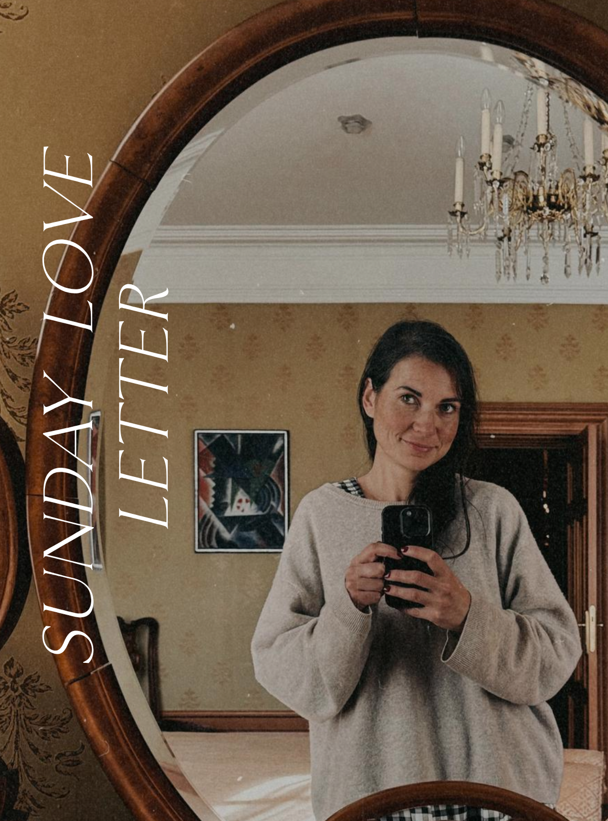 Self-portrait taken in a mirror, showing Elina in a cozy sweater holding her phone, with warm light, vintage details, and a chandelier reflected behind her.