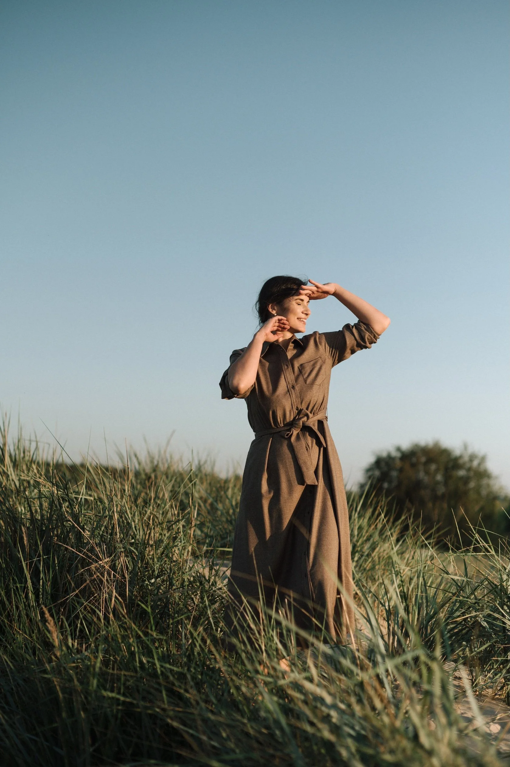 Elina standing among tall beach grasses in a long brown belted dress, smiling with her eyes closed and shielding her face from the golden evening sun beneath a clear blue sky.