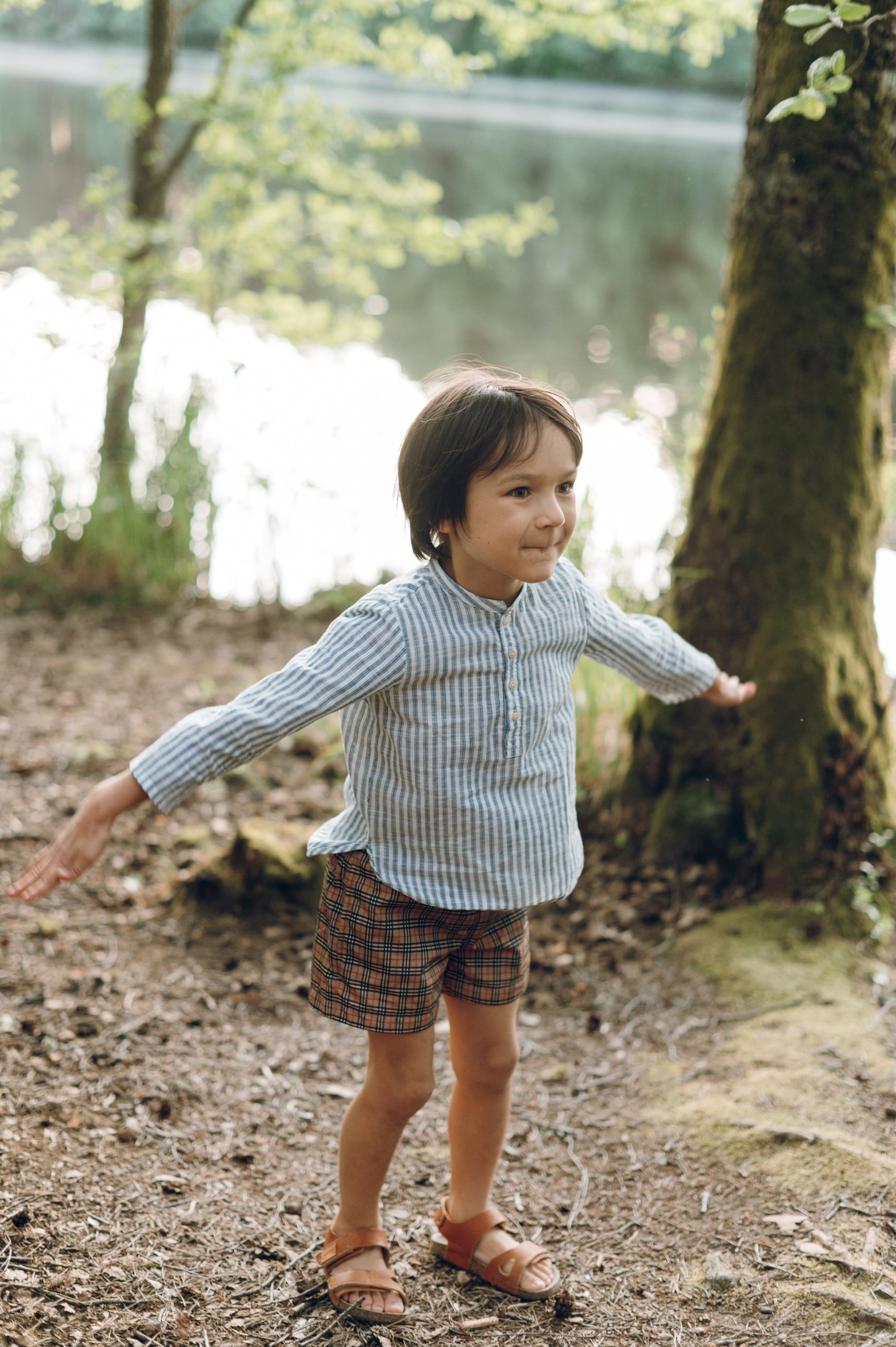 A young child wearing a blue striped shirt, plaid shorts, and brown sandals stands on a woodland path with arms outstretched, surrounded by trees beside a peaceful lake on a sunny day.