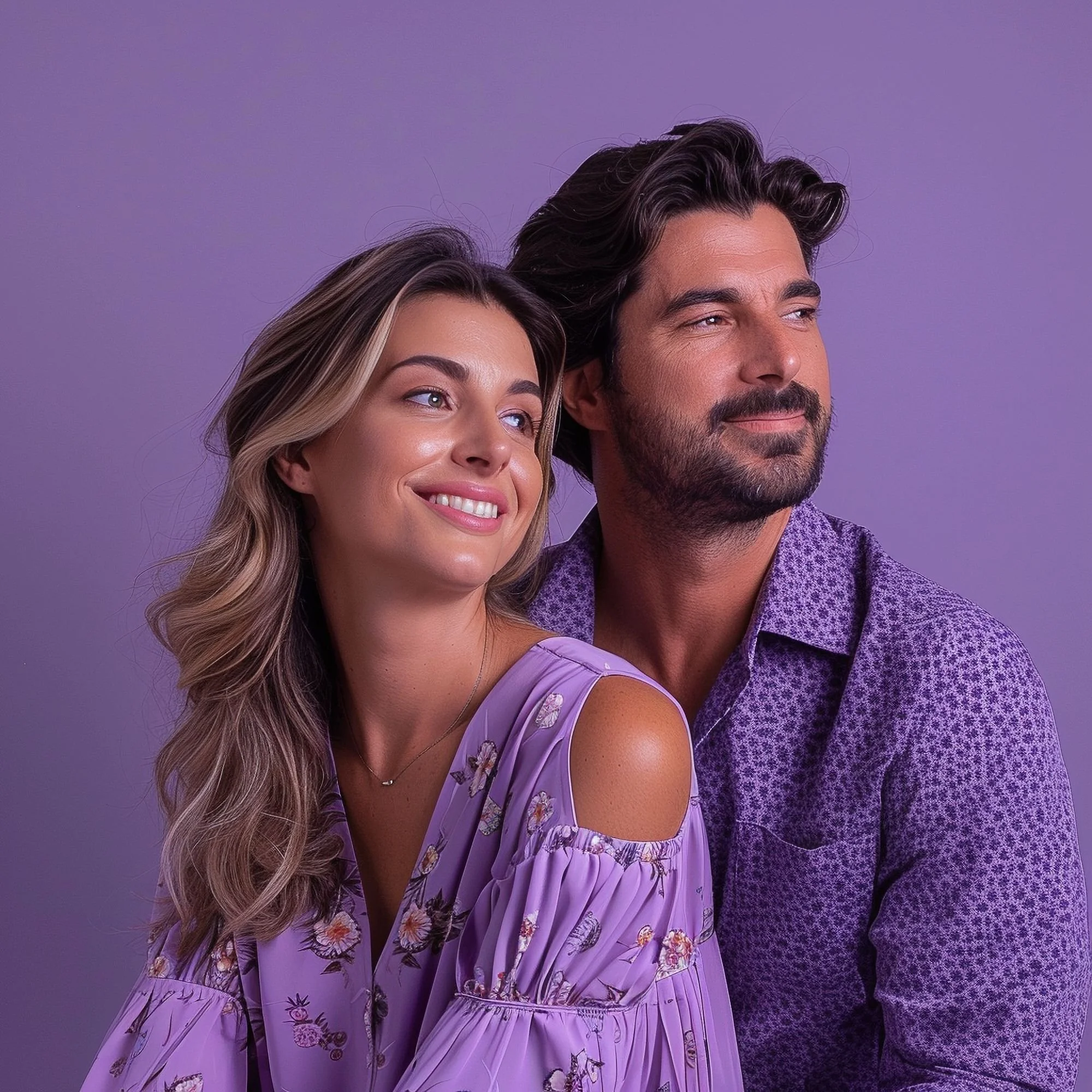 Close-up of a smiling woman with wavy blonde hair and a man with dark, wavy hair and a beard, both looking slightly upward, against a purple background.
