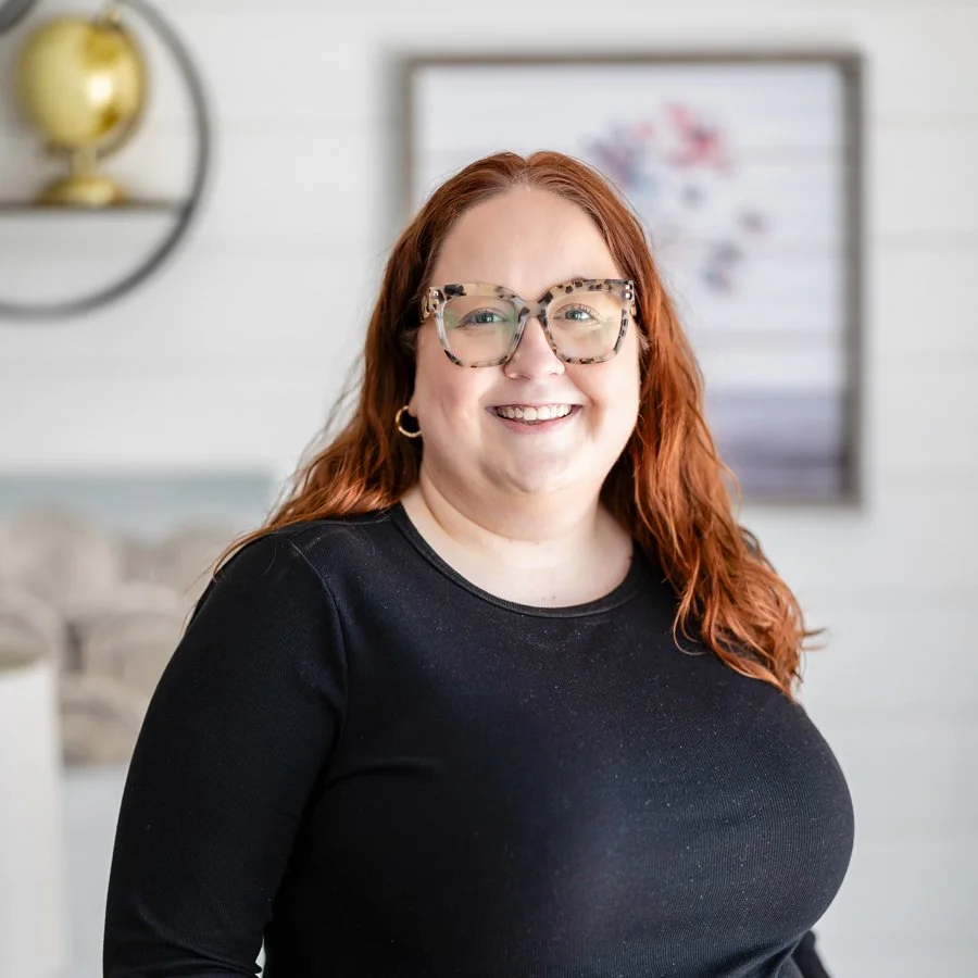 Rachel Galiher, a smiling woman with red hair, glasses, and hoop earrings, wearing a black top, standing indoors with blurred wall art and shelves in the background.