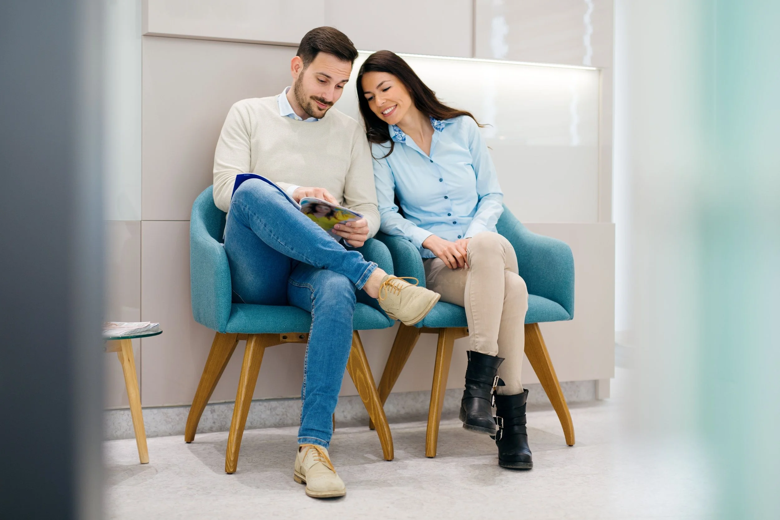 A man and woman sitting close together on a blue chair in a modern, well-lit waiting area, looking at a magazine.