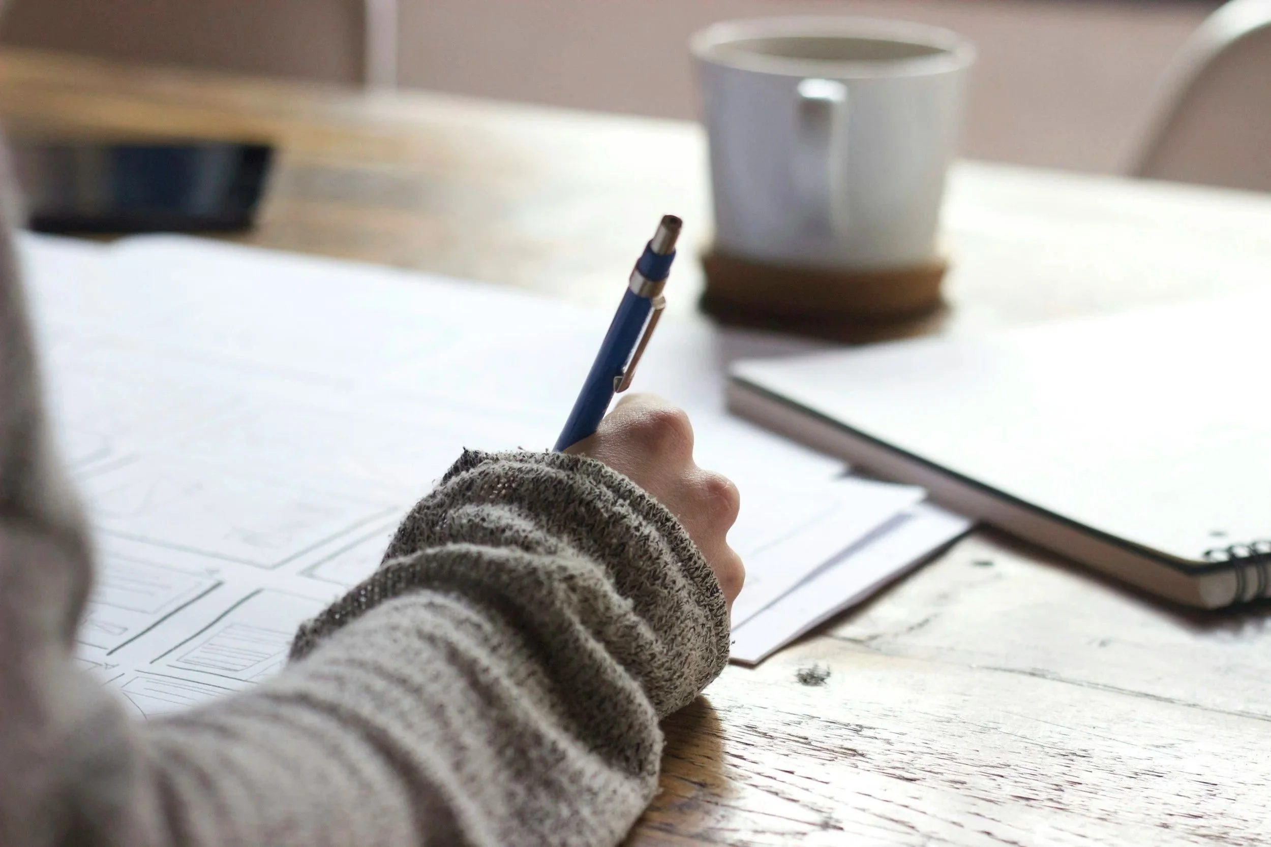 A hand holding a pen over a sheet of paper with diagrams, on a wooden table with a coffee mug, a laptop, and a stack of books or notebooks nearby.