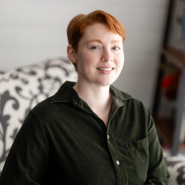 A young woman with red hair, blue eyes, and freckles, smiling, wearing a dark green collared shirt and silver stud earrings, indoors against a plain wall background.