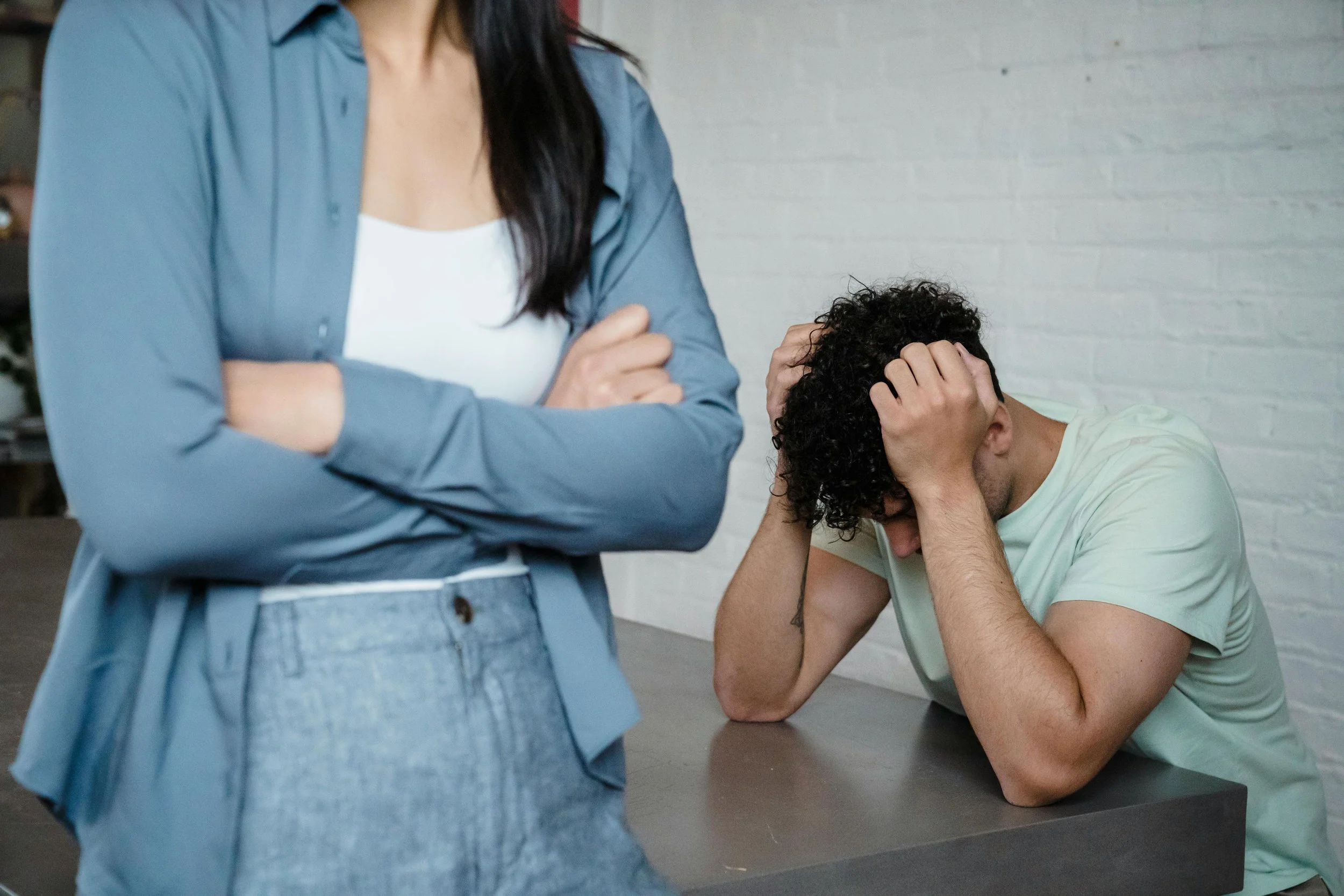 A woman standing with arms crossed and a man sitting at a table with head in hands, appearing distressed or overwhelmed.