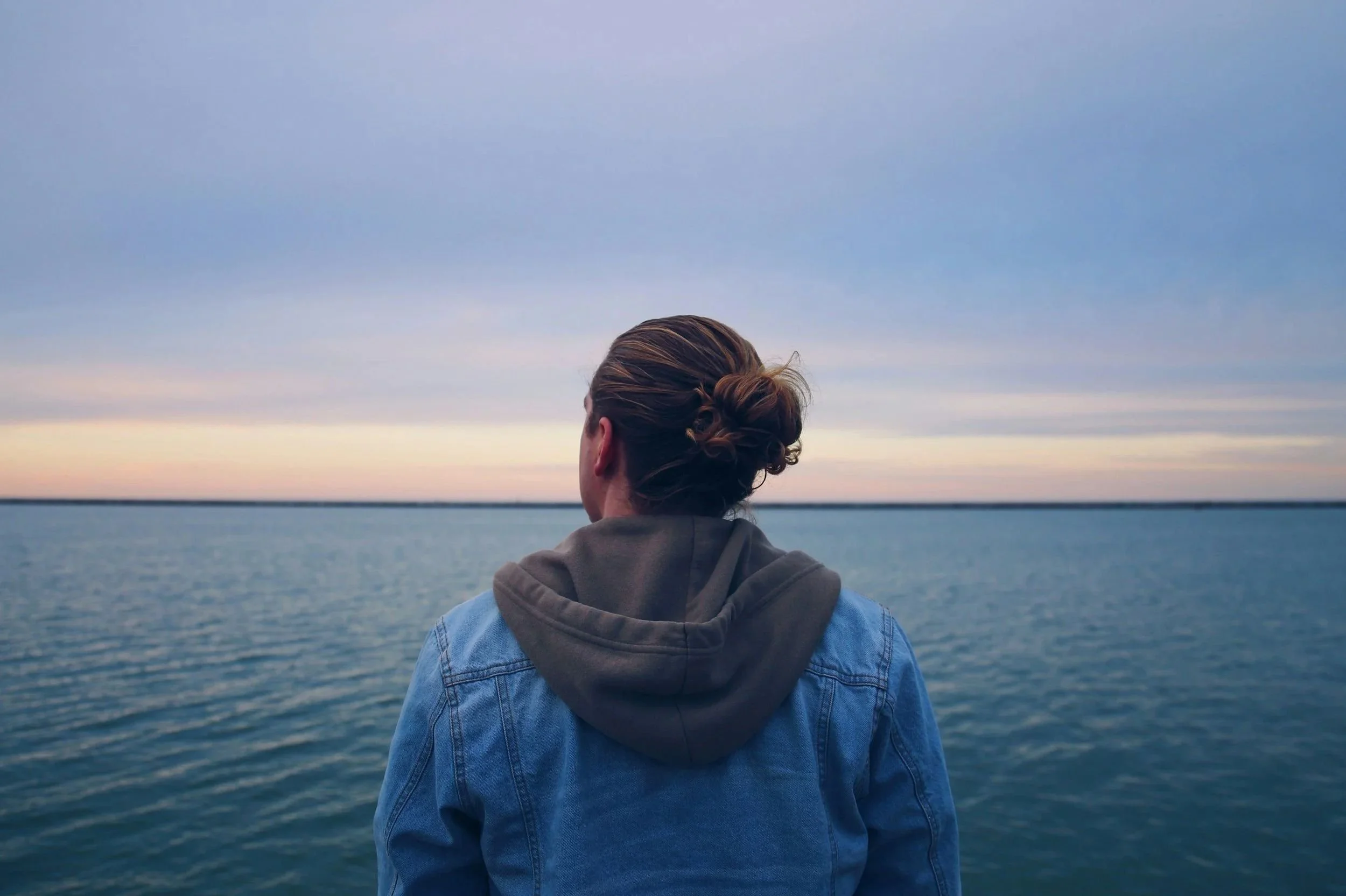A person with brown hair tied in a bun, wearing a denim jacket over a hoodie, standing by a large body of water during sunset or sunrise, gazing at the horizon.