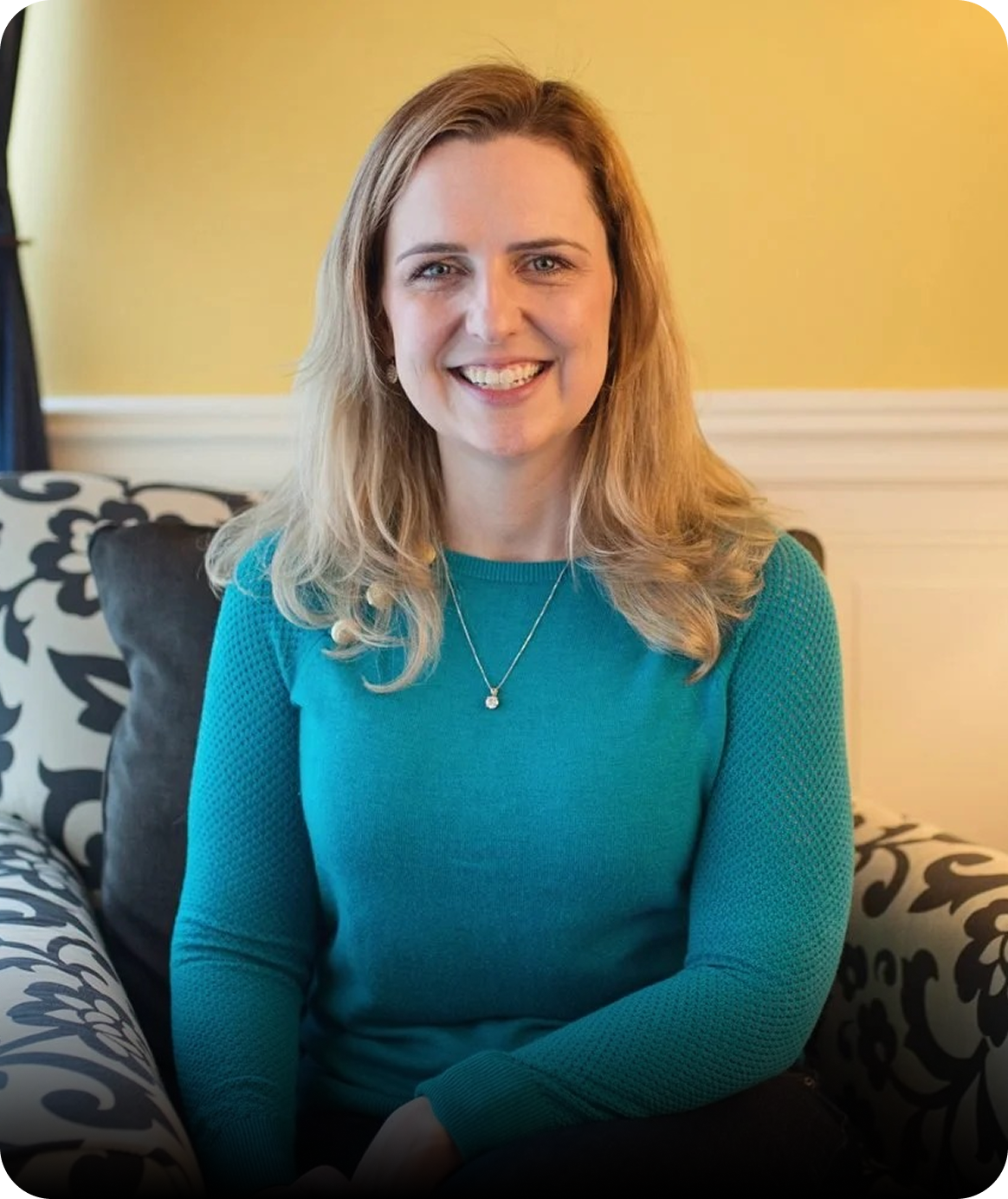 Sarah Olson, LCSW smiling, sitting on a patterned sofa in a yellow-walled room, wearing a teal long-sleeve shirt and a necklace.