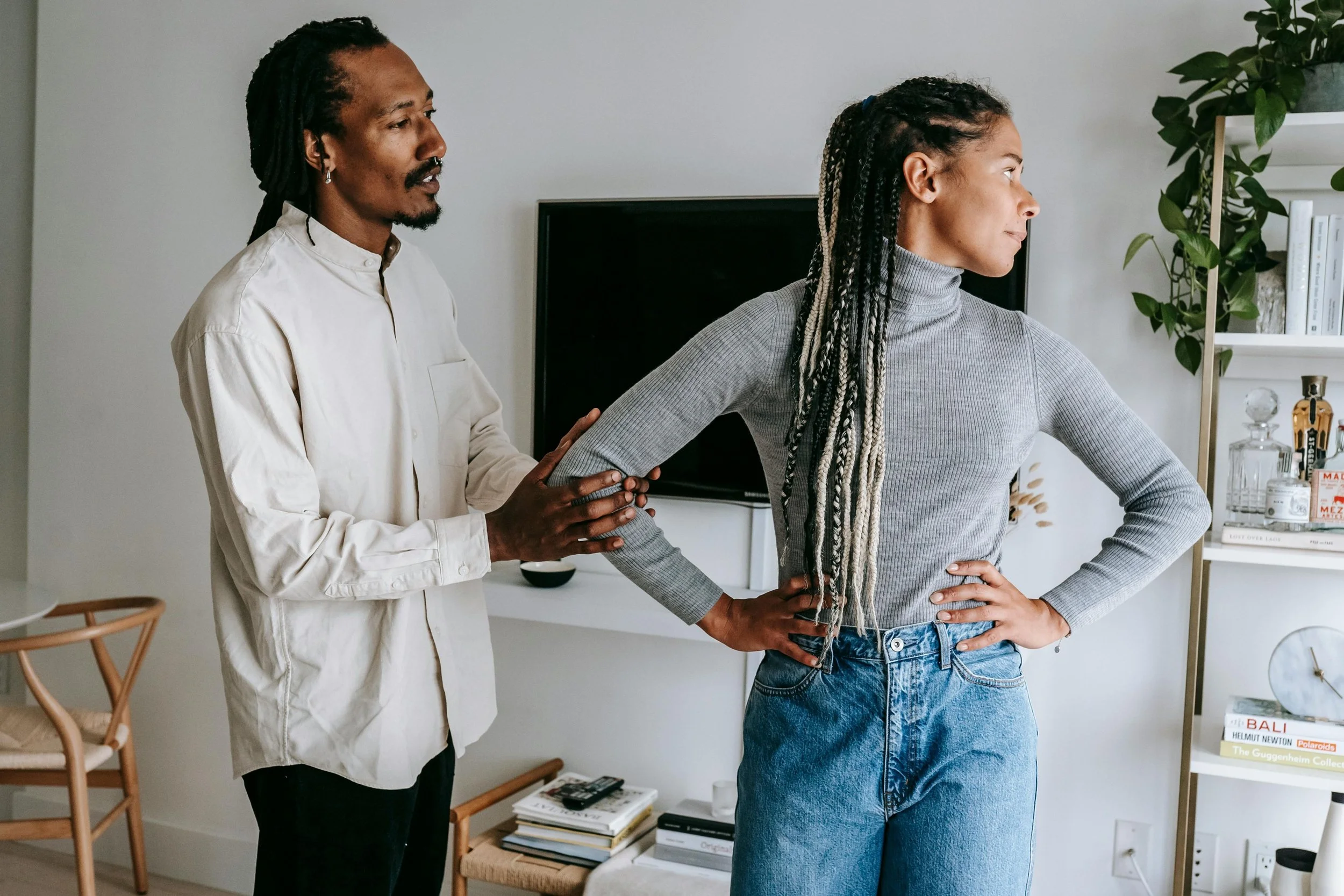 A man with dreadlocks and a woman with braided hair stand in a room, engaging in a conversation or activity. The woman hands on her hip, and the man holds her arm gently while speaking.