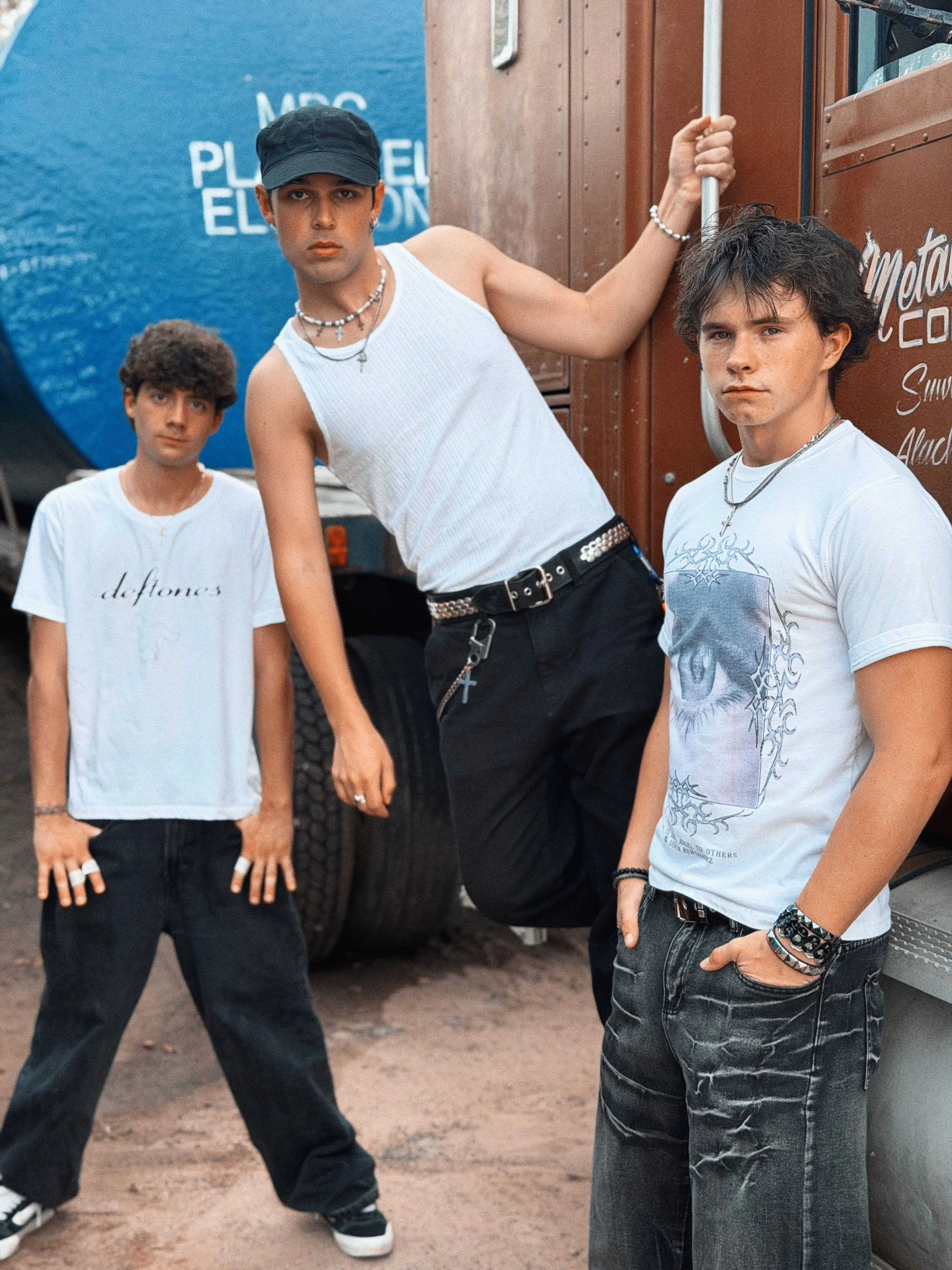 Three young men standing outdoors in front of a truck and blue wall with white text. They are dressed in casual streetwear, with one wearing a black cap, rings, and necklaces. They have different hair styles and serious expressions.