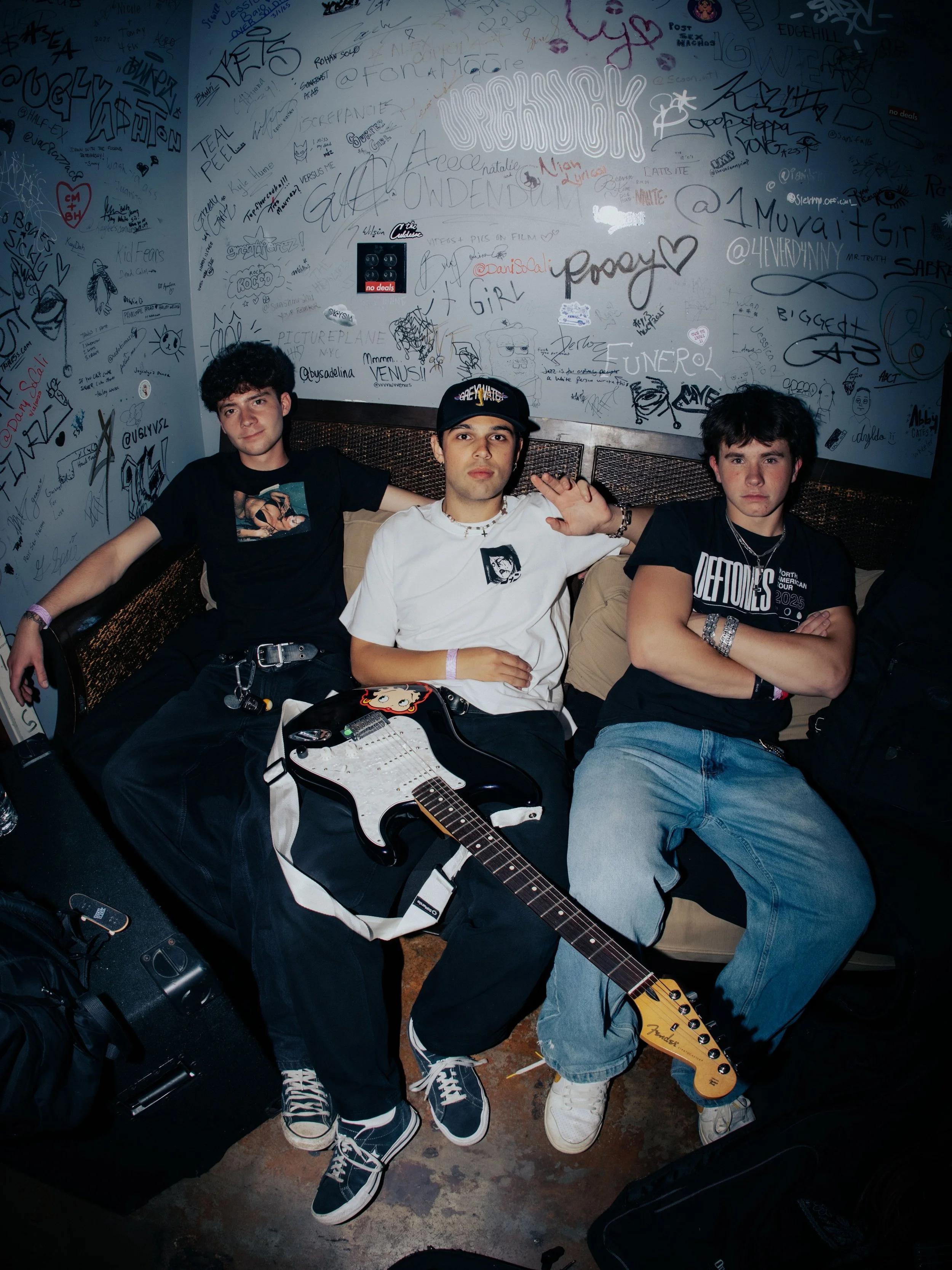 Three young men sit on a bench in a room with graffiti-covered walls. The man in the middle wears a black cap and white T-shirt with a graphic, the man on the right wears a black T-shirt with white text and silver jewelry, and the man on the left has