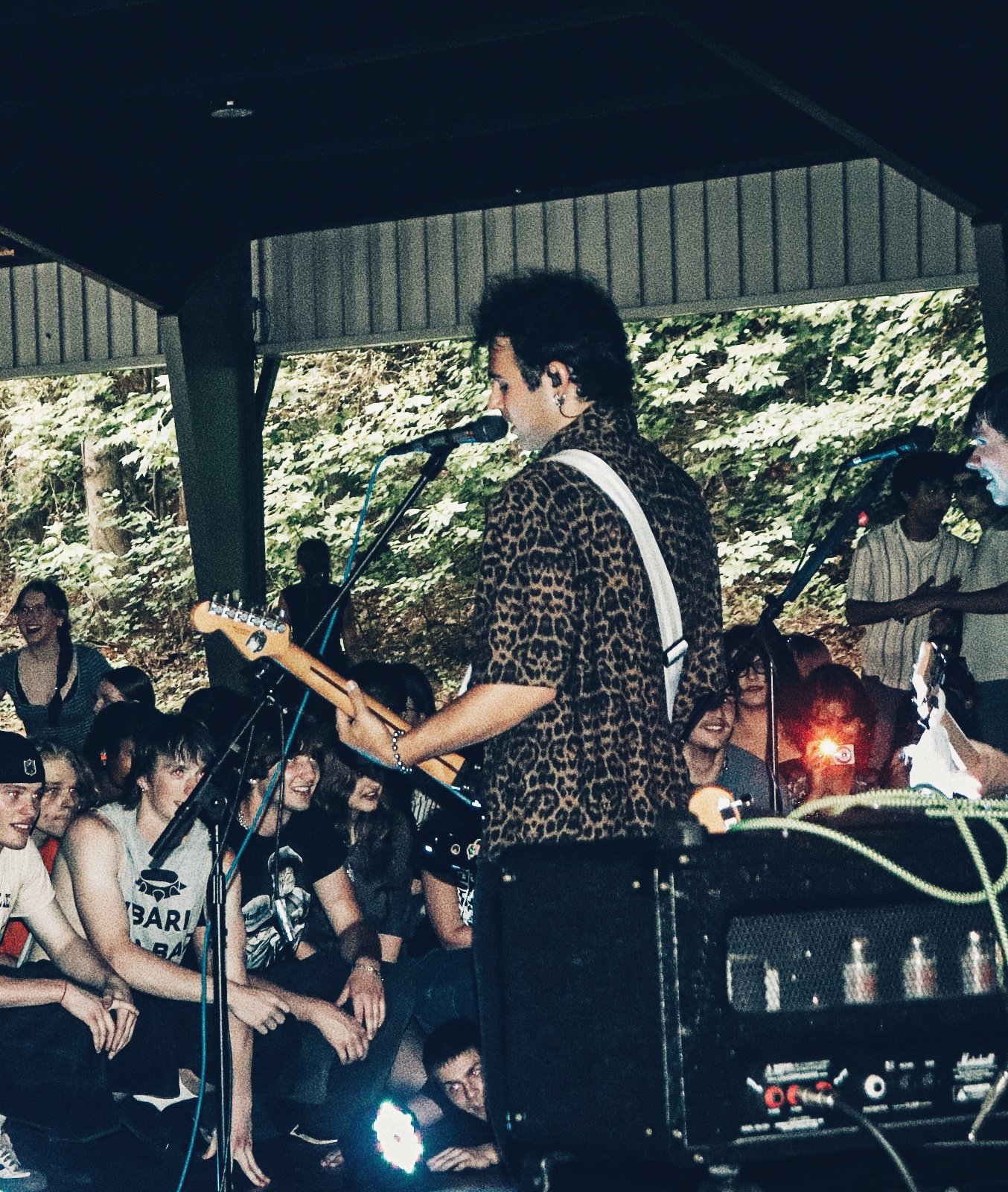 A musician with dark hair, wearing a leopard print shirt, playing an electric guitar and singing into a microphone during a live performance. The background shows an outdoor setting with green trees and an audience smiling and watching the performanc