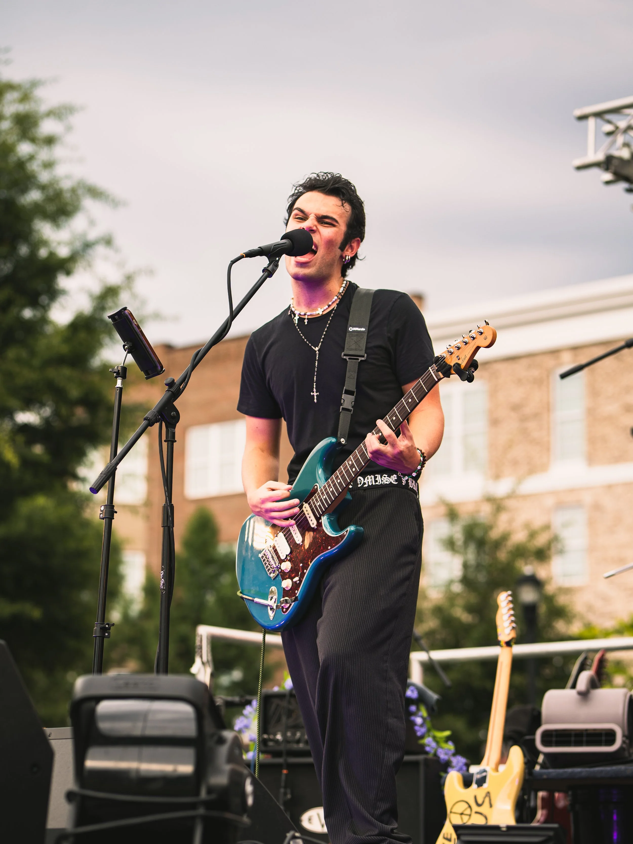 A young man with dark, curly hair and earrings, singing into a microphone while playing an electric guitar during an outdoor performance. He is wearing a black t-shirt, layered necklaces, and dark pants. There are musical equipment and a musical inst