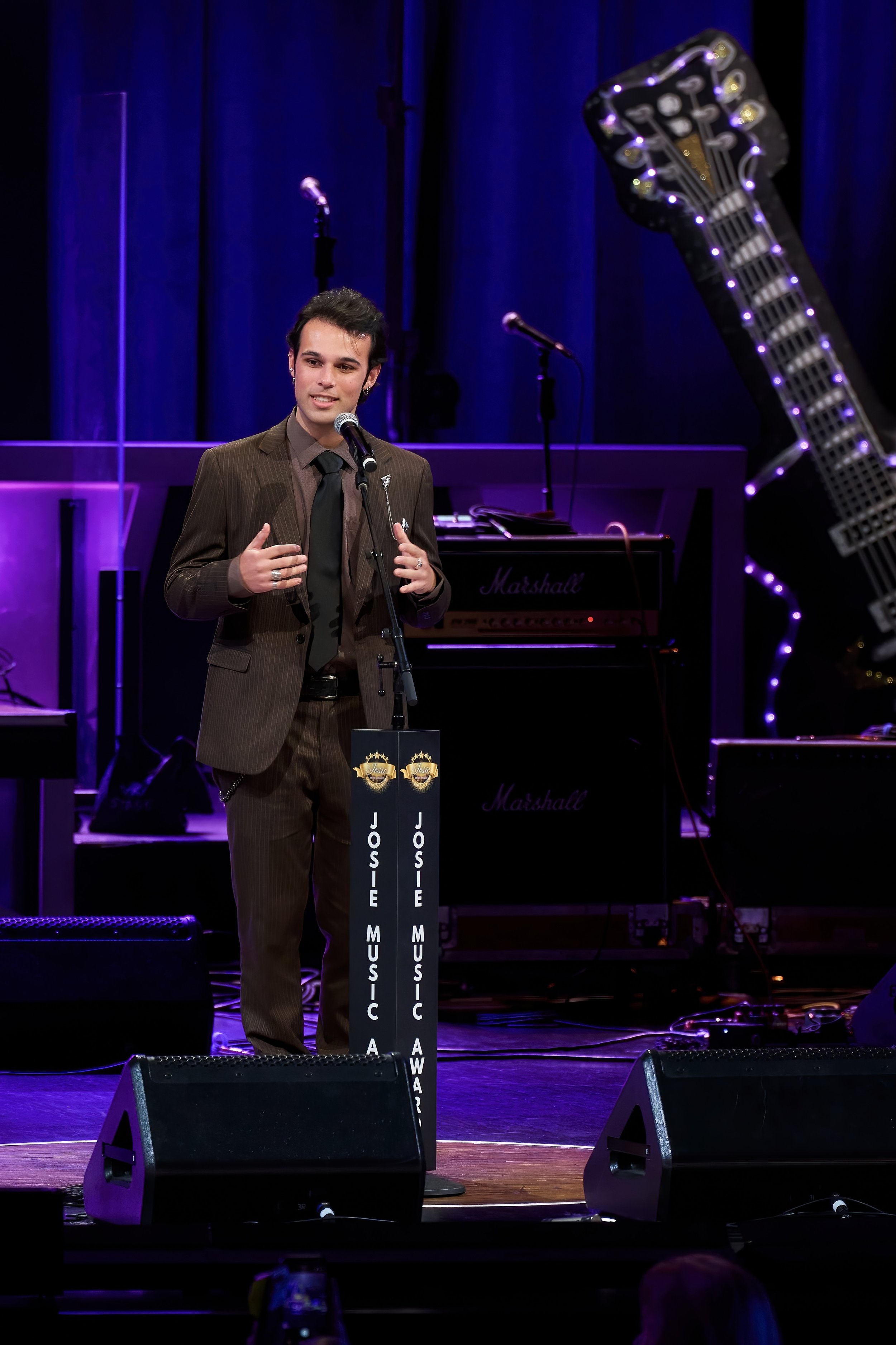 A man in a brown suit and black tie speaking at a music awards event on stage, with a microphone, musical instruments, and purple lighting.