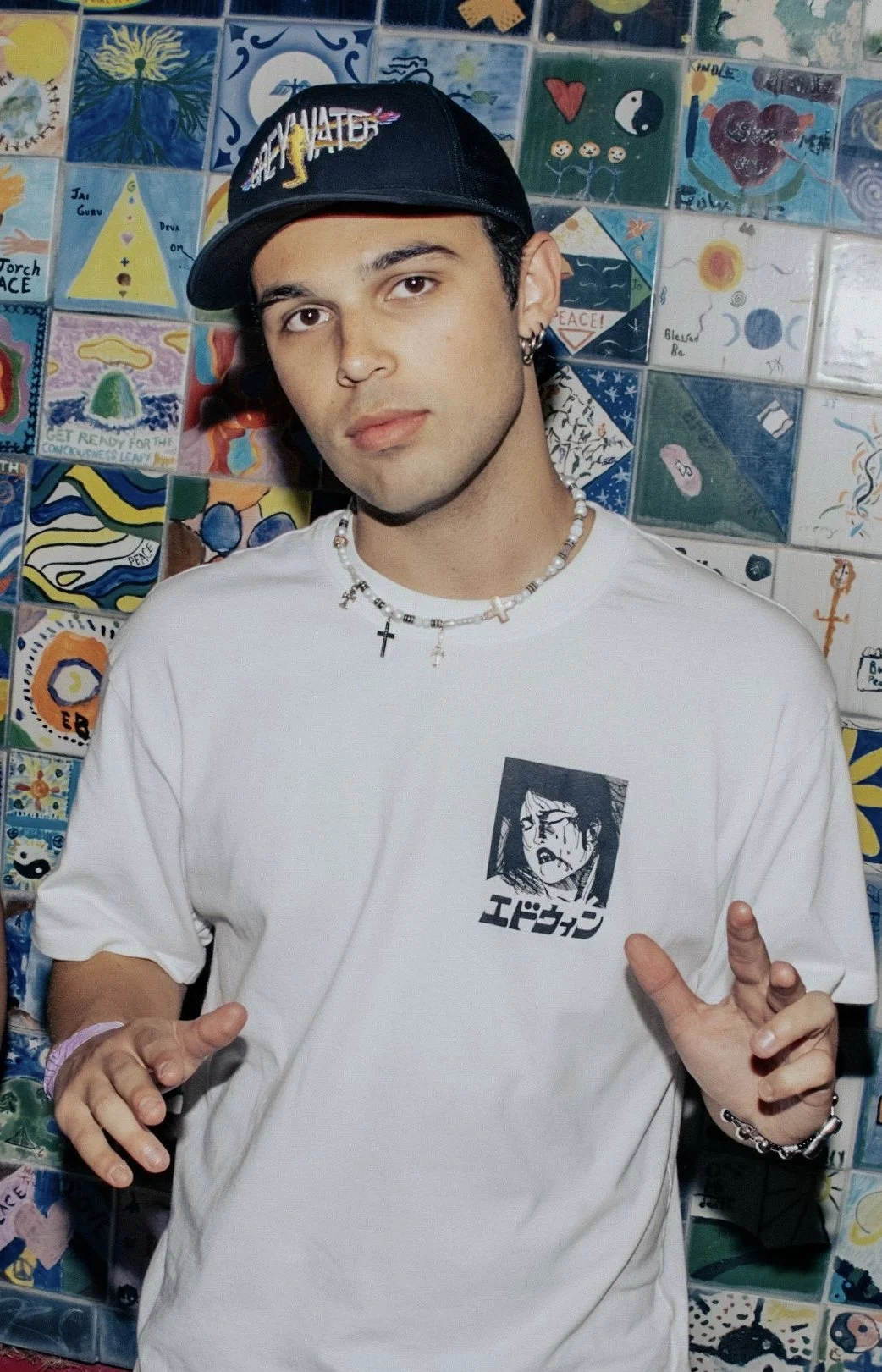 A young man with earrings and a cross necklace wearing a white T-shirt with a black graphic and Japanese characters, black cap, and bracelets, standing in front of a colorful tiled wall with various paintings and drawings.