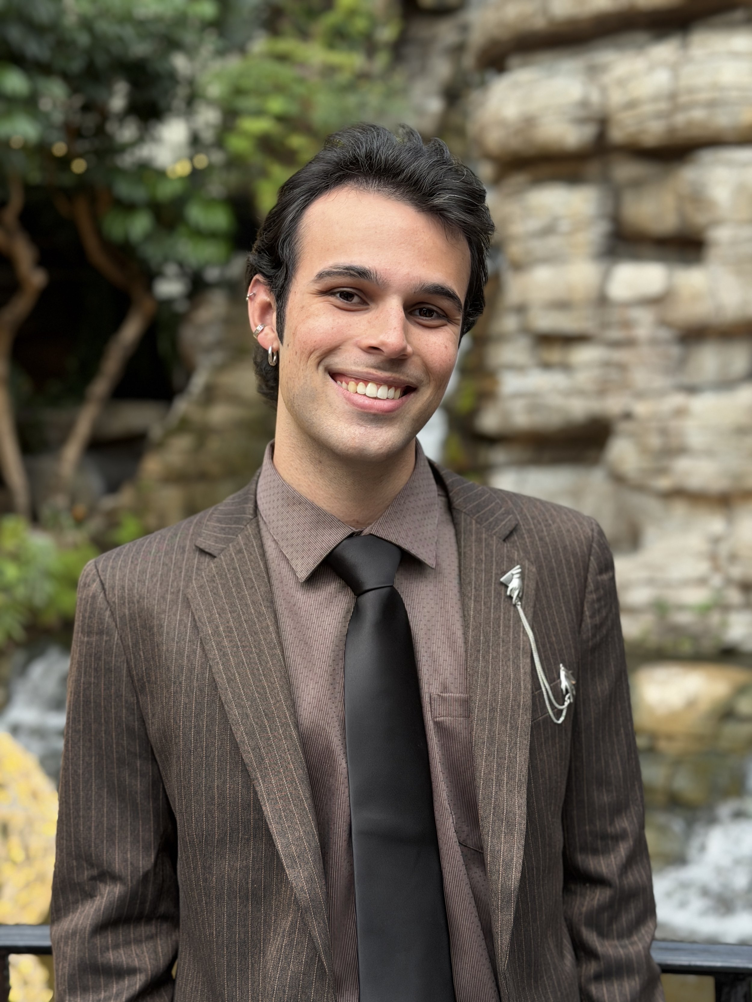A young man with dark hair and earrings smiling outdoors in front of a rocky waterfall and greenery.