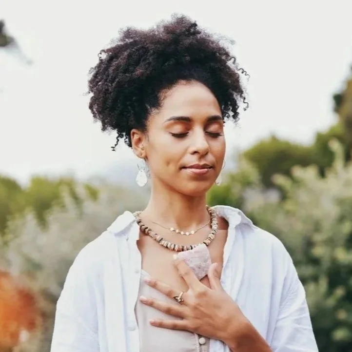 A woman with natural curly hair practicing yoga outdoors with eyes closed, hand on chest, wearing jewelry and a white shirt.