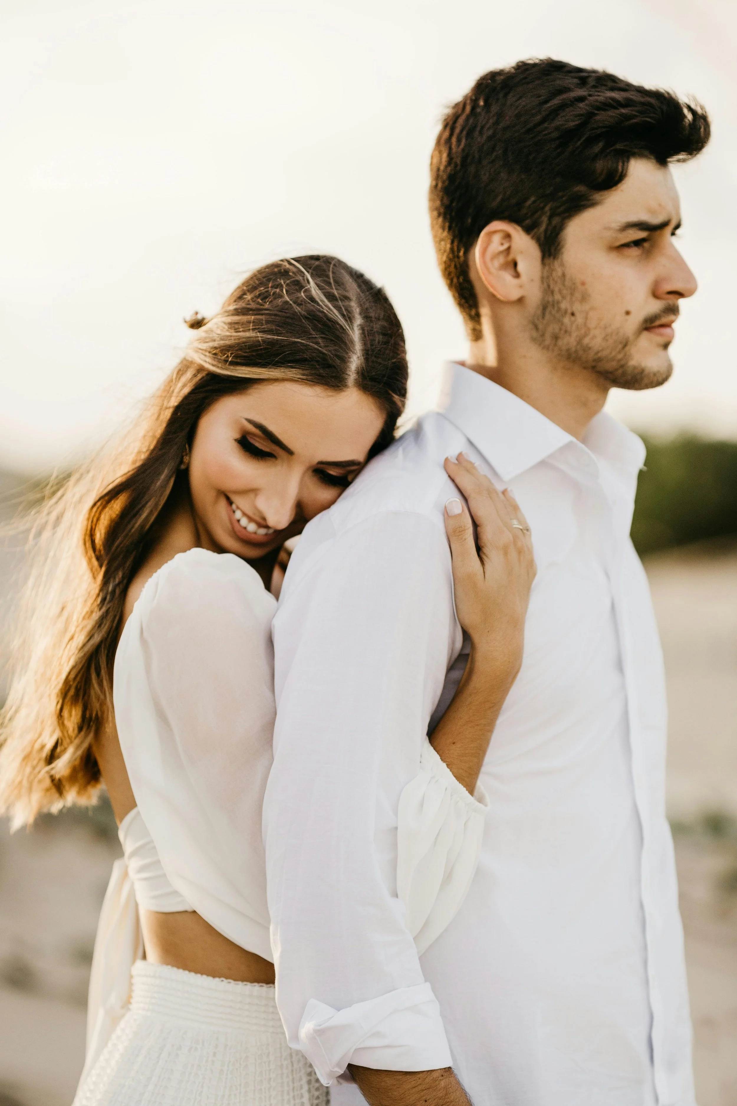 A woman leans on a man's back with her eyes closed, smiling, while the man looks away with a serious expression, both dressed in white, outdoors at sunset.
