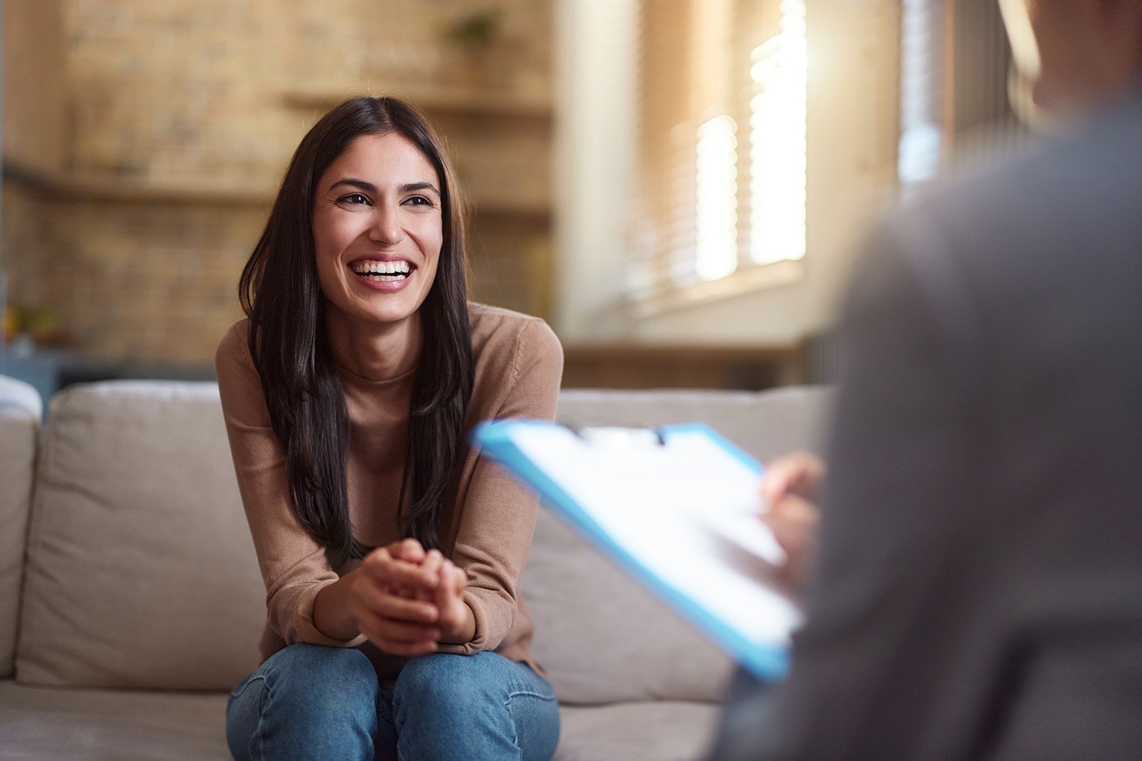 A woman with long dark hair, wearing a beige top, smiling and sitting on a sofa during a conversation, with a person holding a clipboard in front of her.