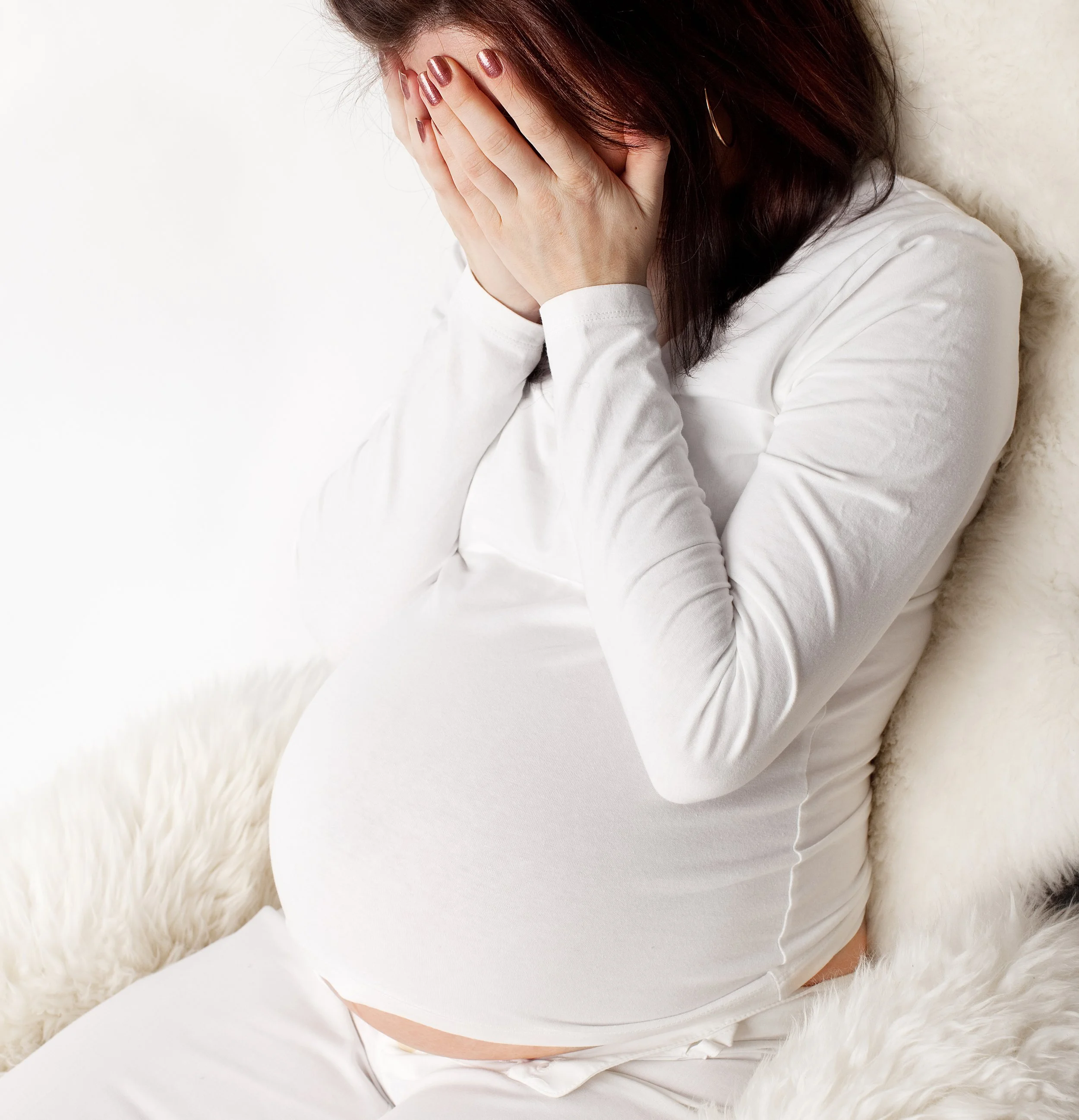 Pregnant woman sitting on a fluffy white surface, covering her face with her hands.