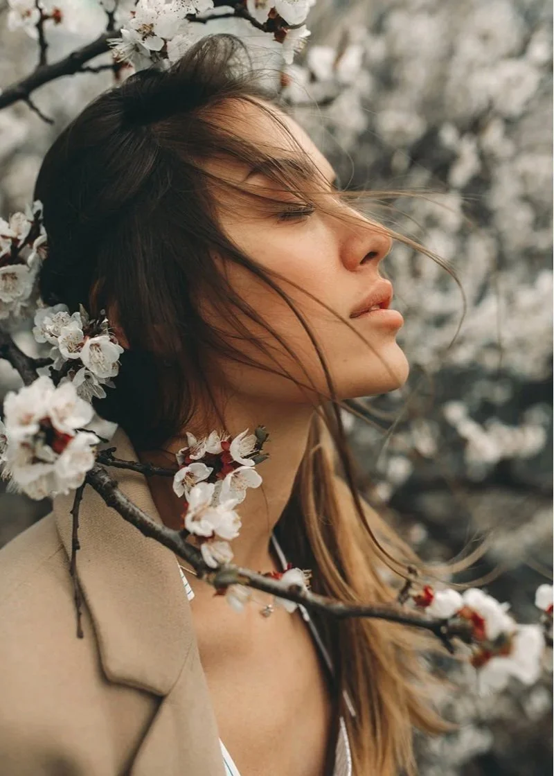 A woman with long brown hair and closed eyes flowers her face as she faces a blooming tree with white flowers.