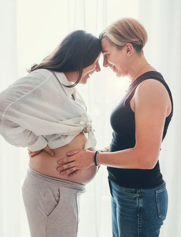 Two women, one pregnant, face each other with foreheads touching, smiling, in front of a bright window.