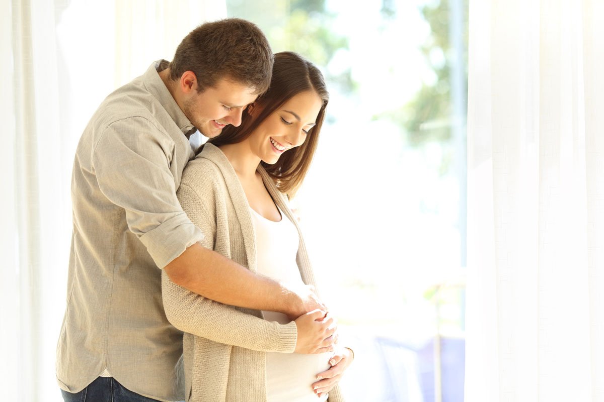 A smiling couple with a pregnant woman holding her belly, standing indoors near a large window with natural light.
