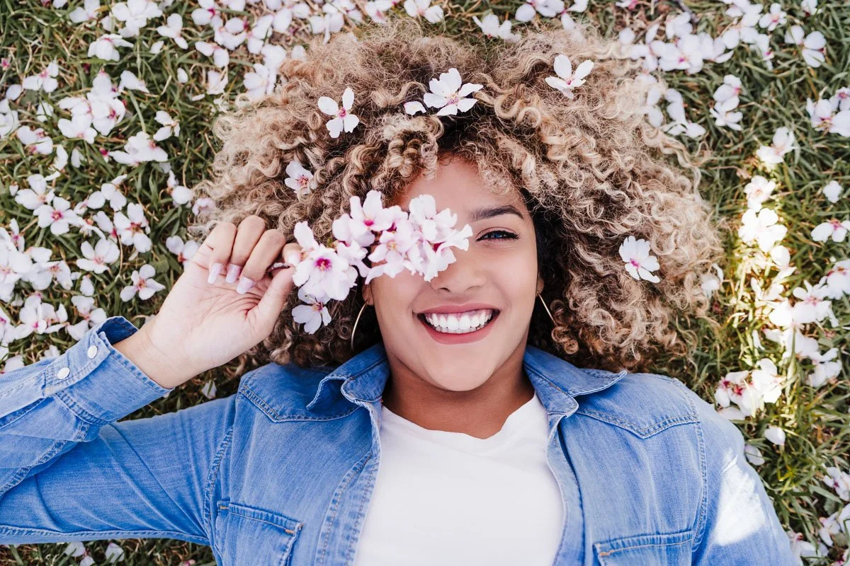 A woman with curly hair lying on a bed of white flowers, holding a bunch of flowers near her face, smiling.