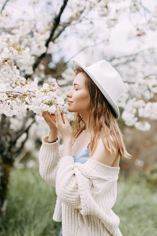 A woman in a white hat and beige knitted cardigan smelling cherry blossoms in a spring garden.