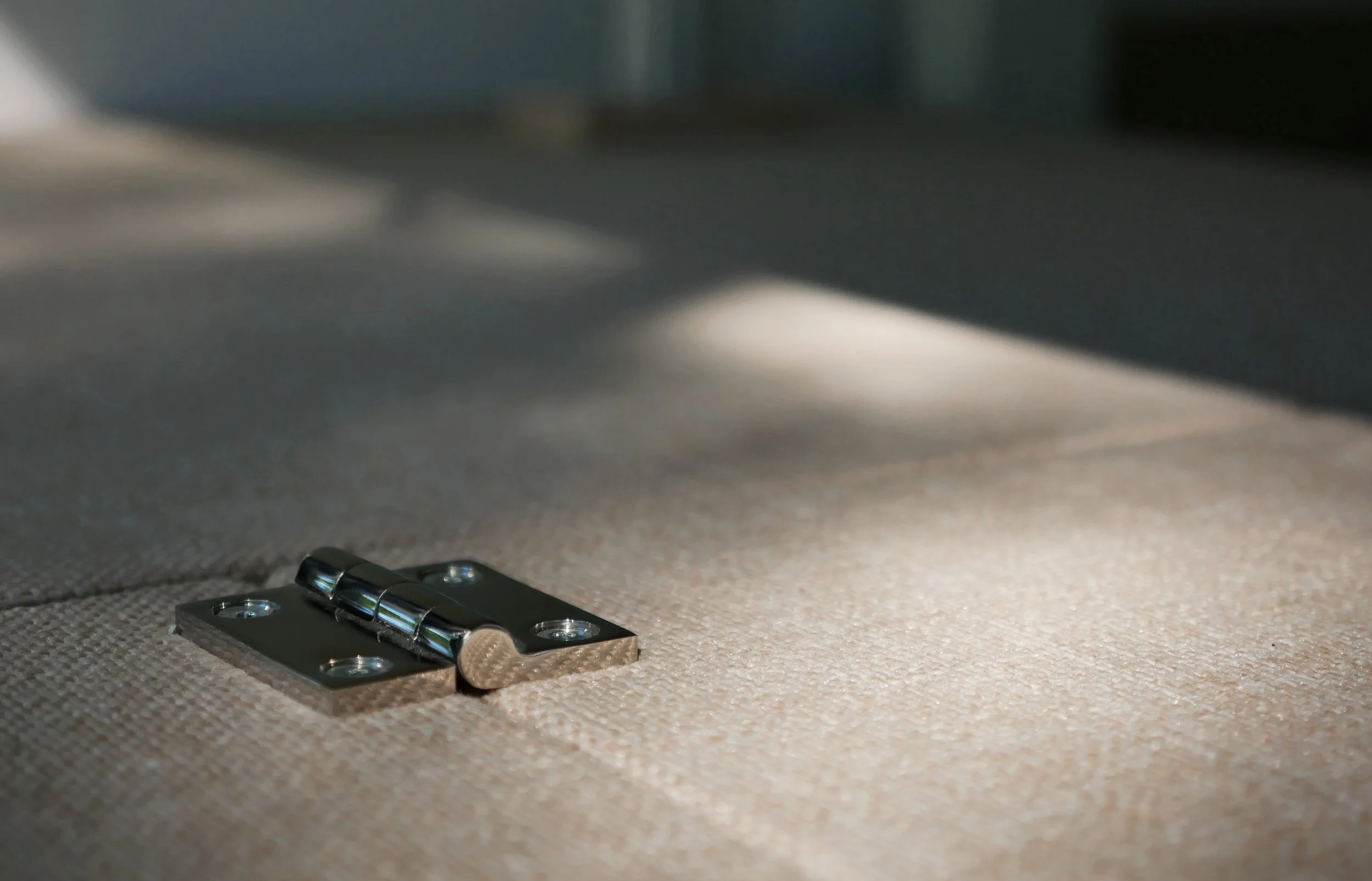 Craftsmanship detail in 'Bjorn' van conversion — macro close-up of a polished chrome butt hinge resting on a natural tweed upholstered panel, highlighting hardware quality and precision, dramatic diagonal light across the woven fabric.