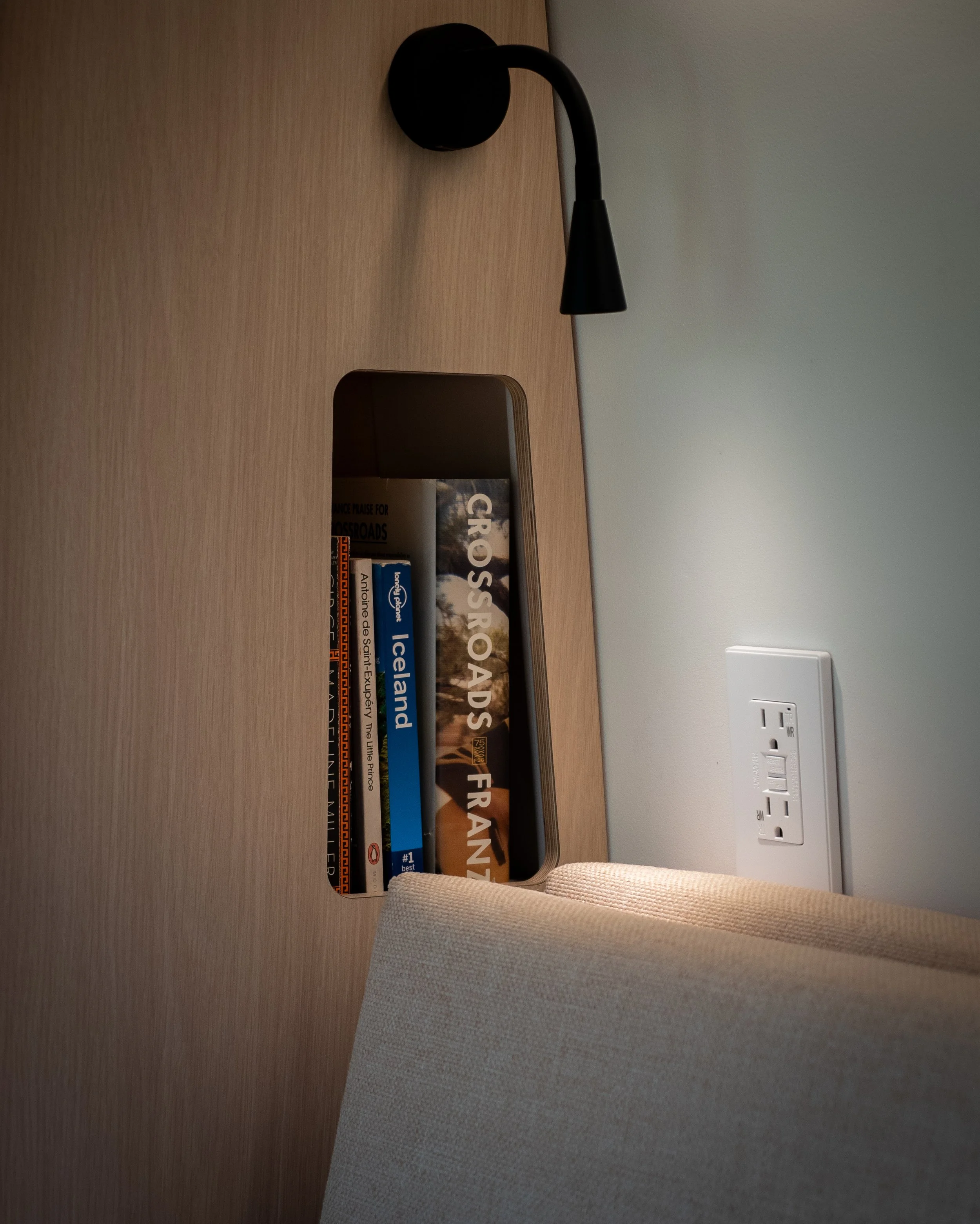Bedside reading nook in 'Aina' van conversion — black wall-mounted gooseneck reading sconce above a recessed birch plywood shelf holding Lonely Planet Iceland, Crossroads, and France travel guides; white 120V outlet to the right, cream cushion with w