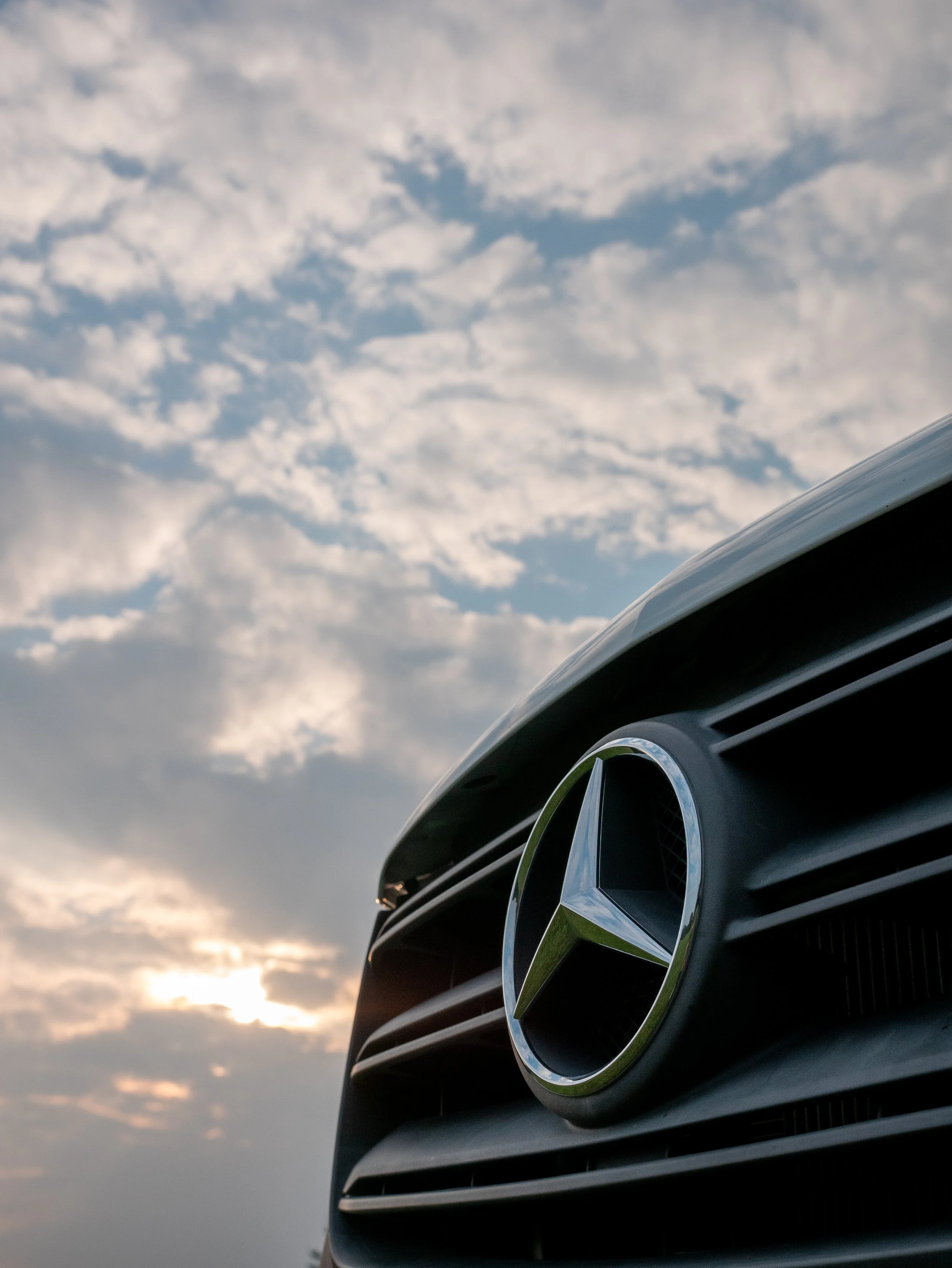 Dramatic exterior detail of 'Aina' Mercedes Sprinter van conversion — chrome three-pointed star grille badge shot from below against a moody sky with scattered clouds and a setting sun behind.
